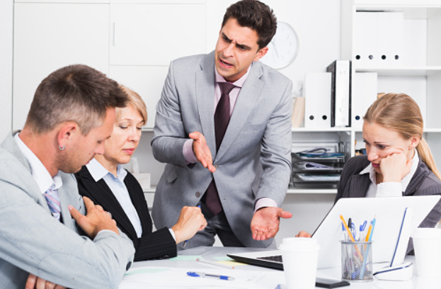 A man in a gray suit is speaking passionately to a group of three colleagues in an office setting, with two women and a man listening attentively and looking contemplative.