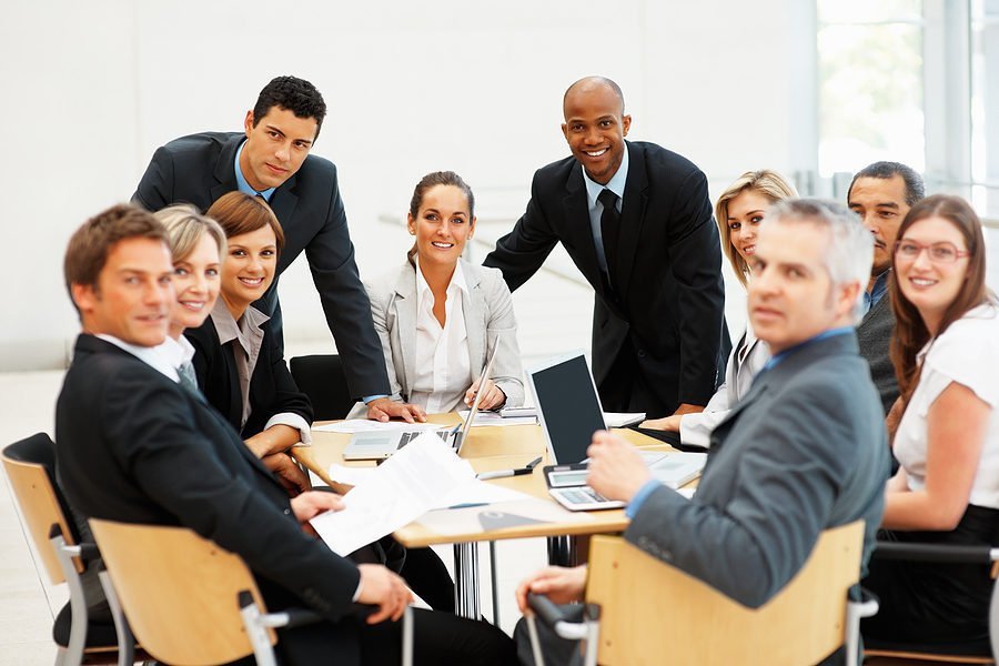 A diverse group of business professionals gathered around a conference table in a modern office, smiling and engaging in a meeting.