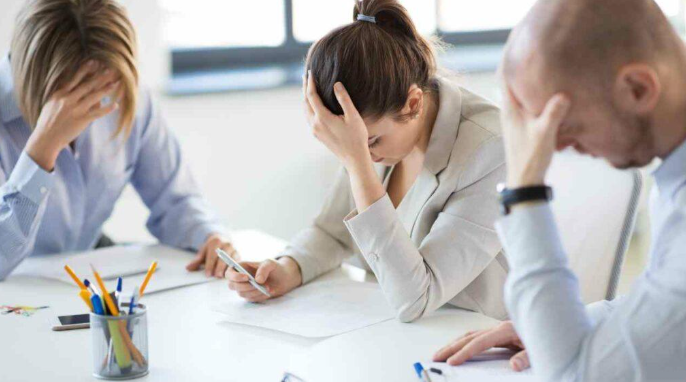 Three coworkers are sitting at a table looking stressed, with their heads in their hands, in an office setting.