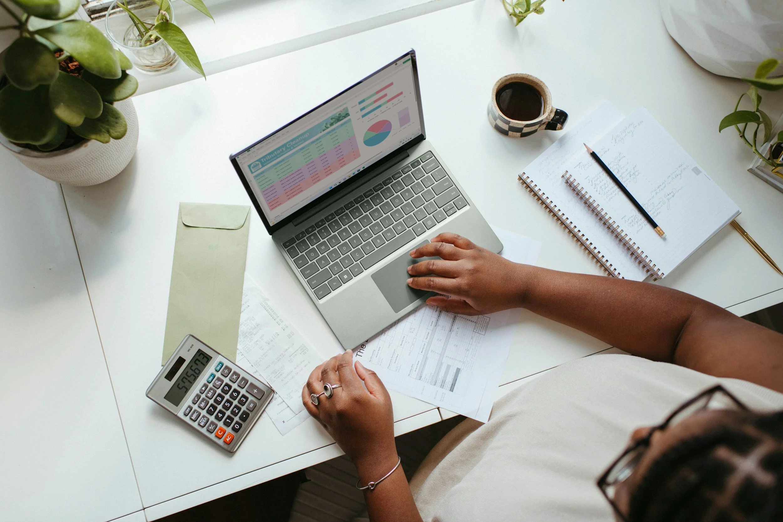 A person working at a desk with a laptop displaying colorful charts and spreadsheets, a coffee mug, a calculator, notebooks, a pen, and some papers.
