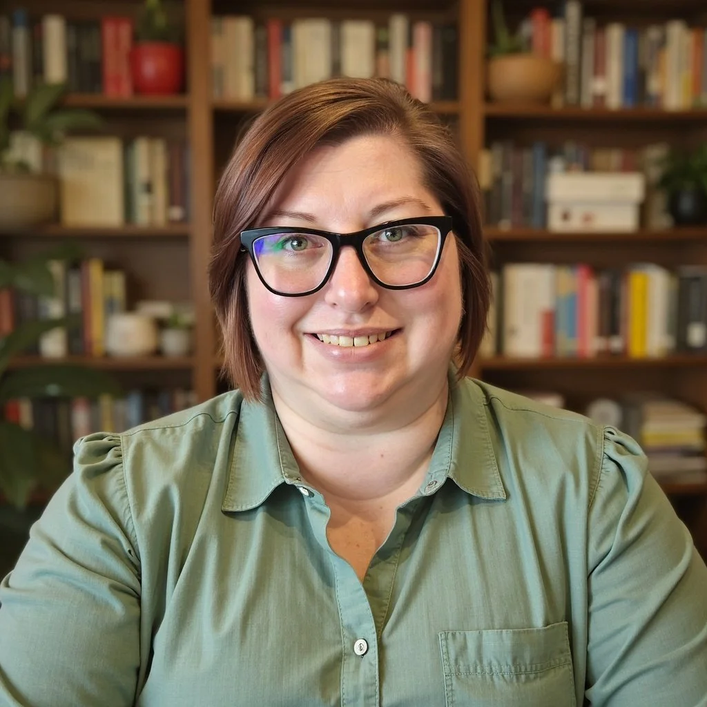A woman with short brown hair, wearing glasses and a green shirt, sitting in front of a bookshelf filled with books and potted plants, smiling at the camera.