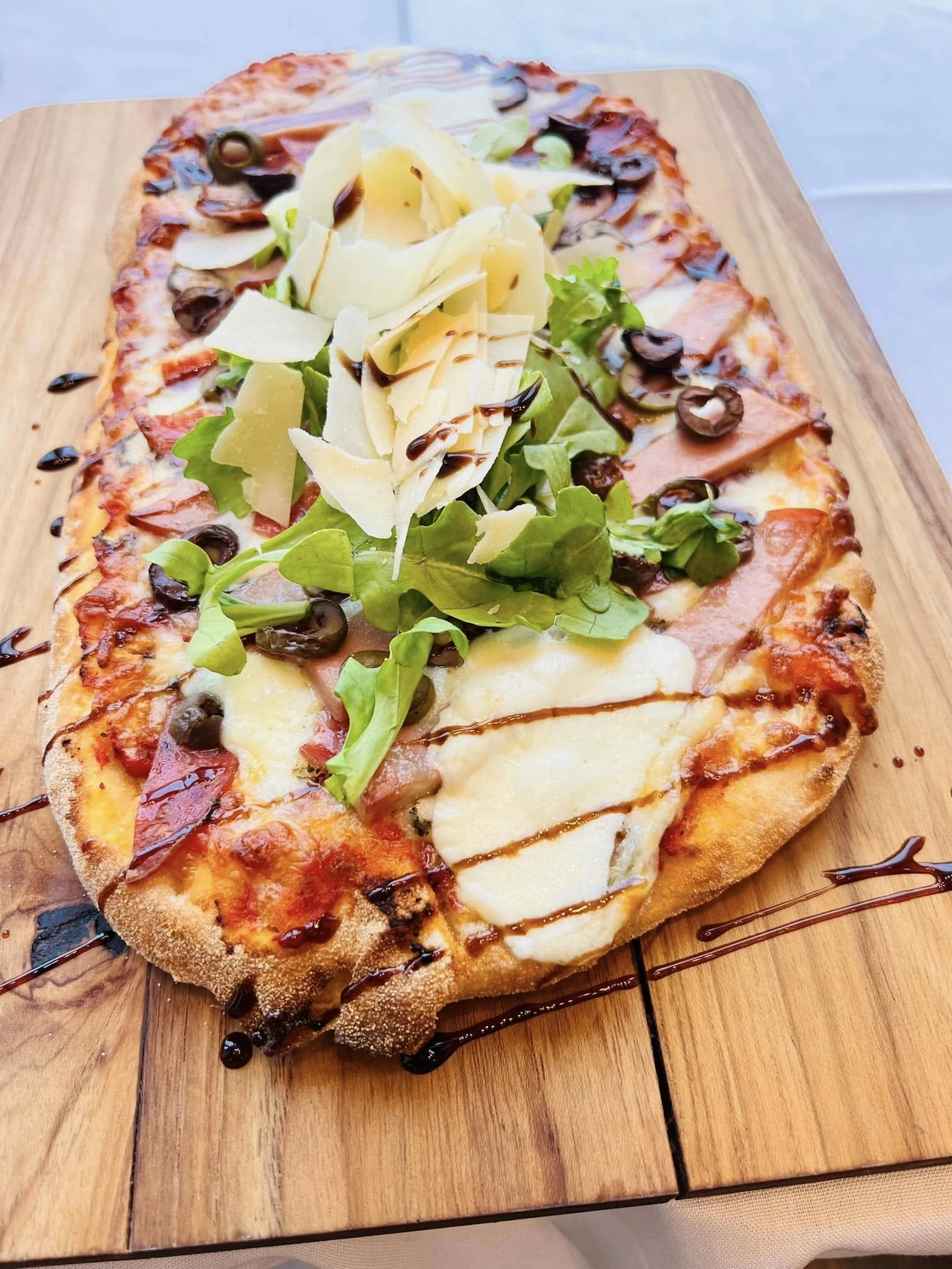 A rectangular pizza with cheese, ham, black olives, lettuce, and cheese shavings, garnished with a drizzle of balsamic glaze on a wooden serving board.