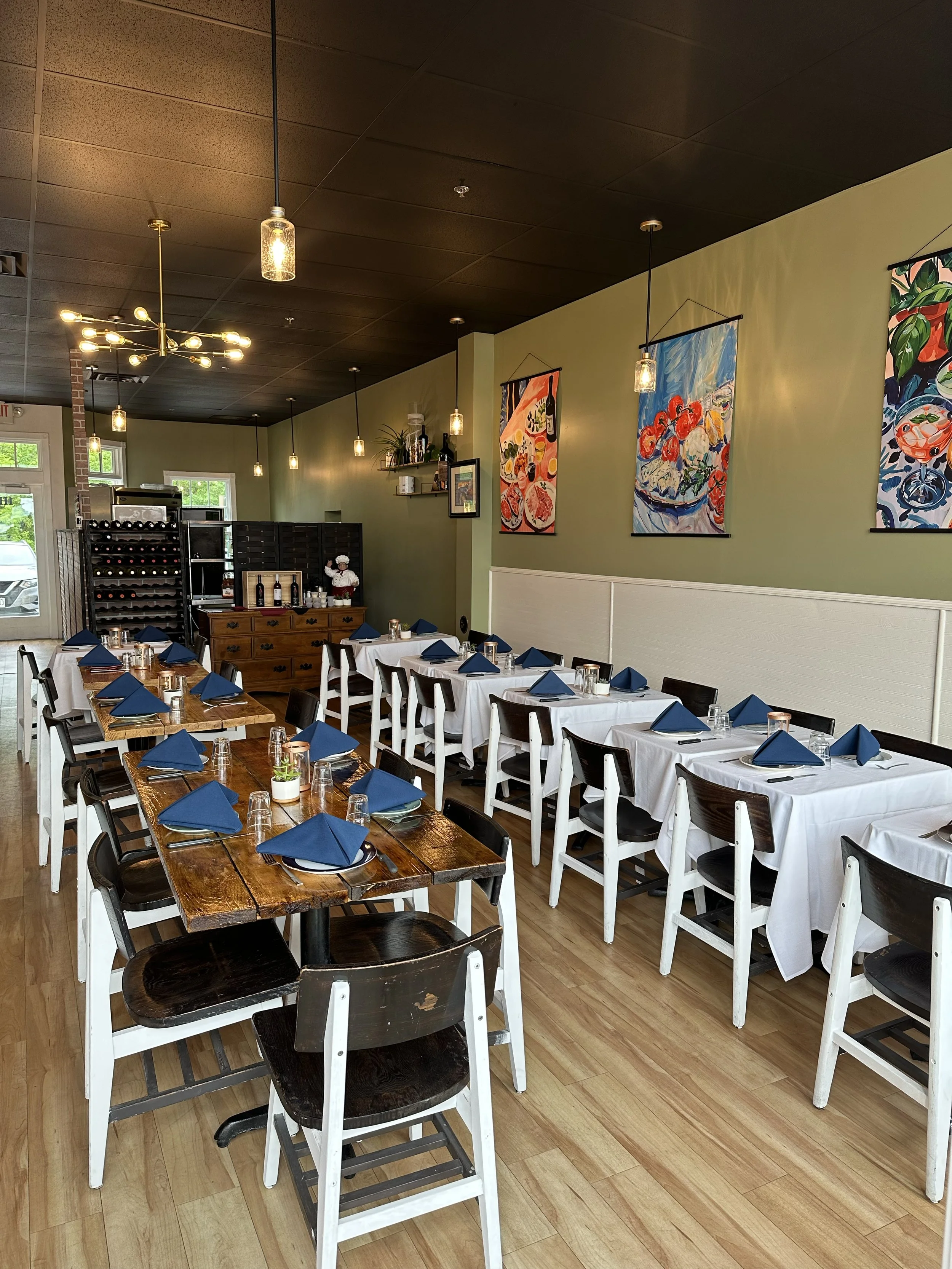 A restaurant dining area with several tables set with white tablecloths, blue napkins, and glassware. The walls feature colorful abstract artwork, and the room is lit with warm hanging pendant lights and a modern chandelier. There are windows allowing natural light.