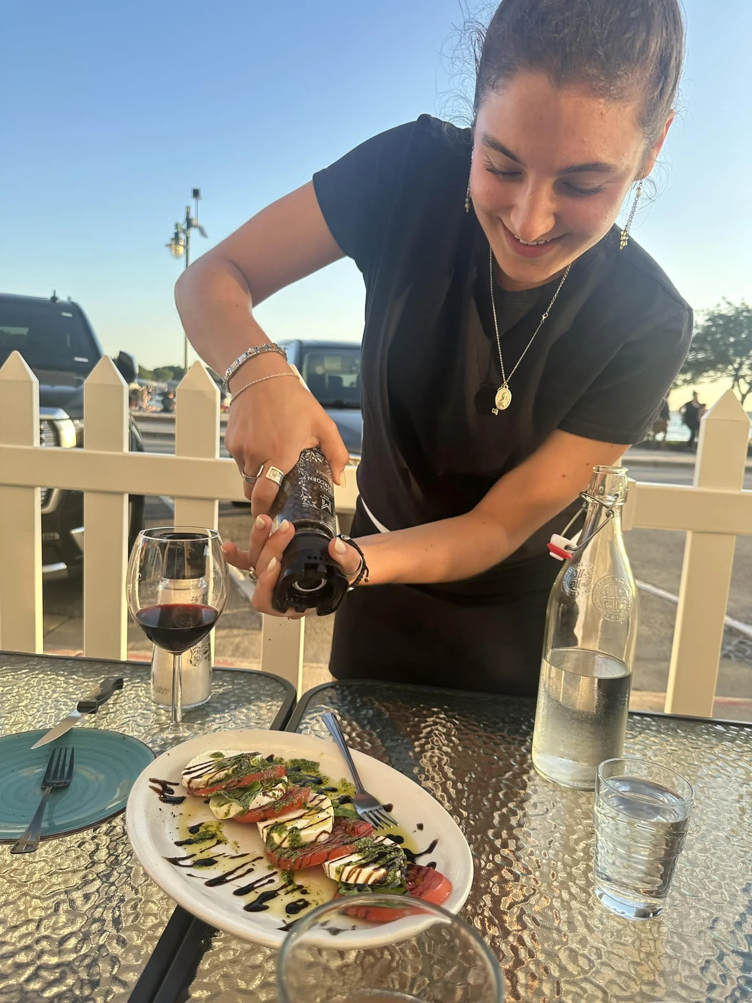 A woman with brown hair tied back, wearing a black top and jewelry, is smiling while pouring a drink at an outdoor dining table during sunset. The table has a platter of sliced vegetables with balsamic drizzle, a glass of red wine, and a bottle of sp