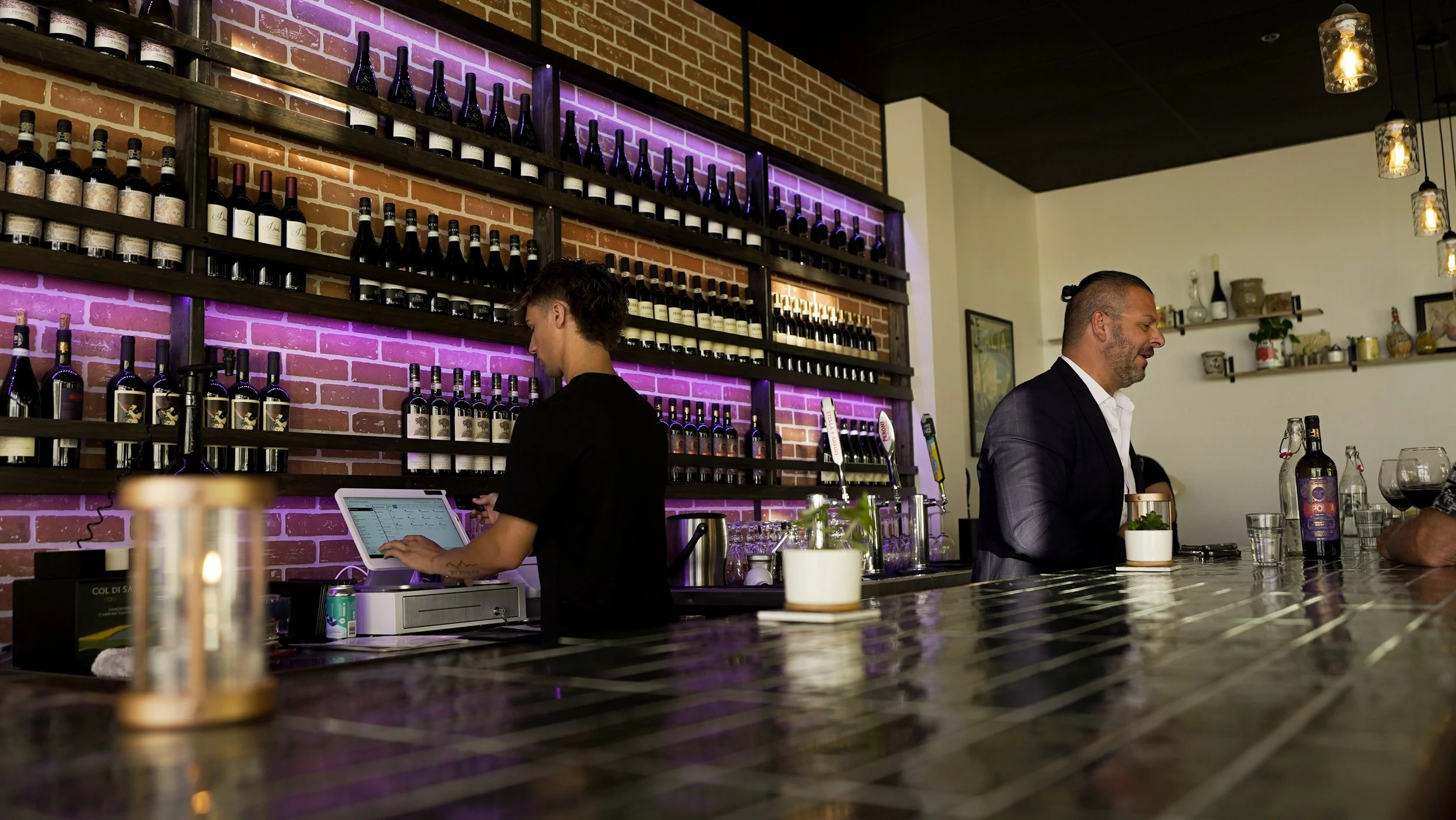Bar scene with a bartender and a customer. The bartender, a woman, is working with a touchscreen register. The customer, a man, is smiling and holding a drink, with bottles of wine and various bar equipment on the counter. Shelves with bottles and a 