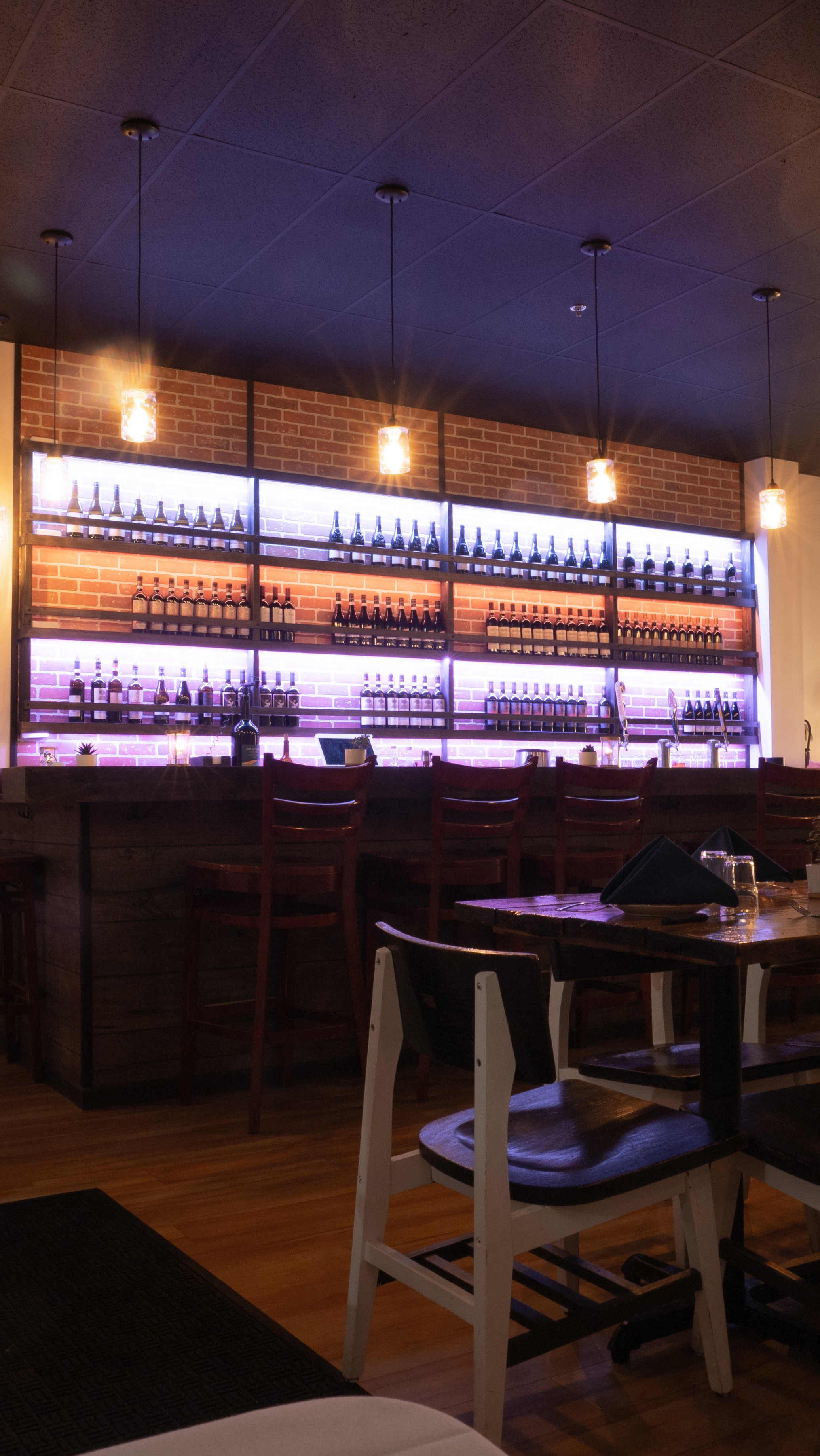 Interior view of a bar or restaurant with a brick wall, illuminated shelves with bottles, pendant lights, and wooden tables and chairs.