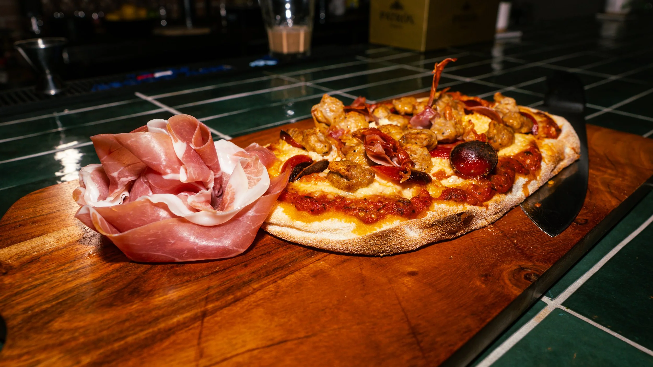 A slice of pizza with various toppings including sausage, pepperoni, and black olives on a wooden serving board, accompanied by a small pile of prosciutto.