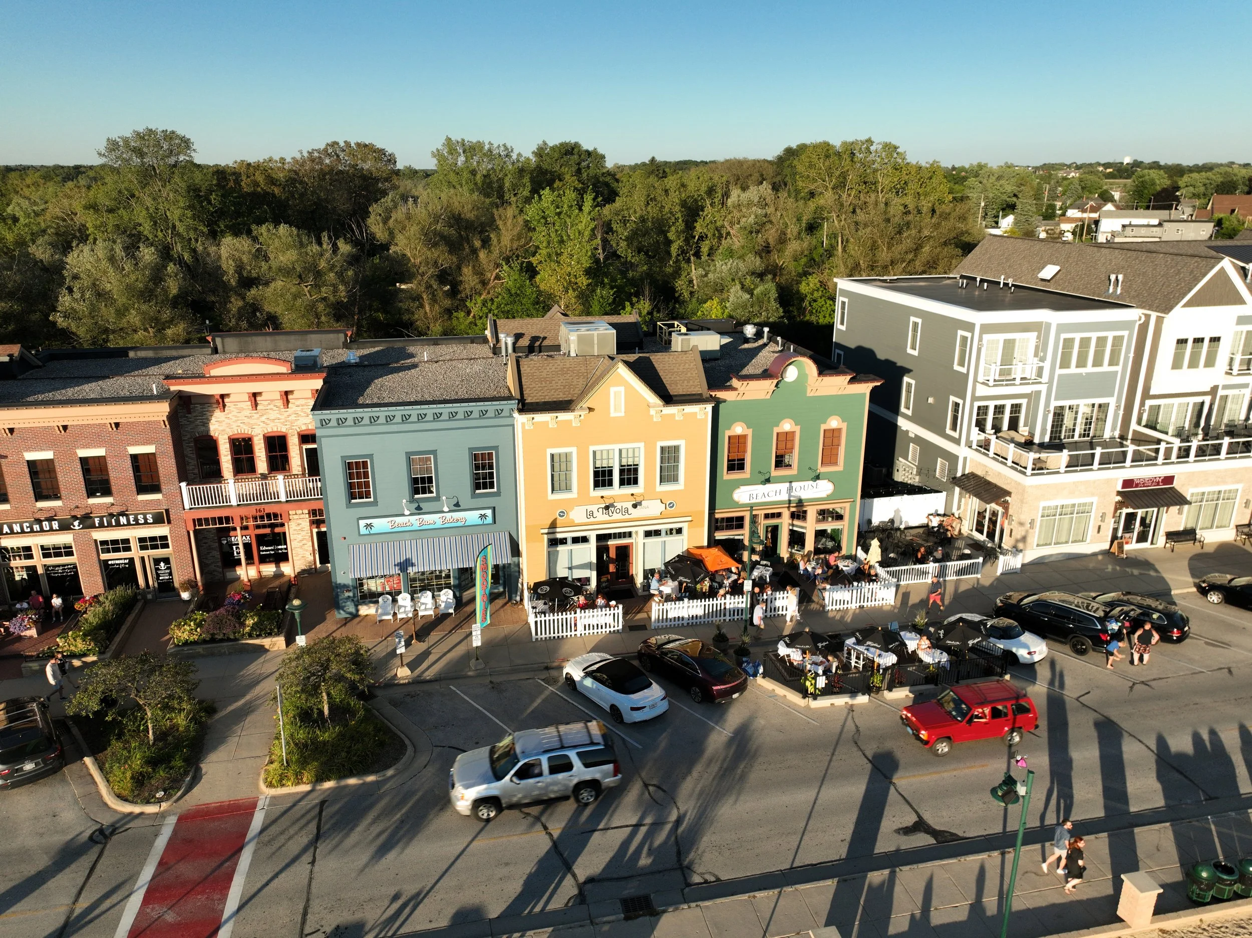 Aerial view of colorful shops on a street with outdoor dining and parked cars, with trees and greenery in the background.