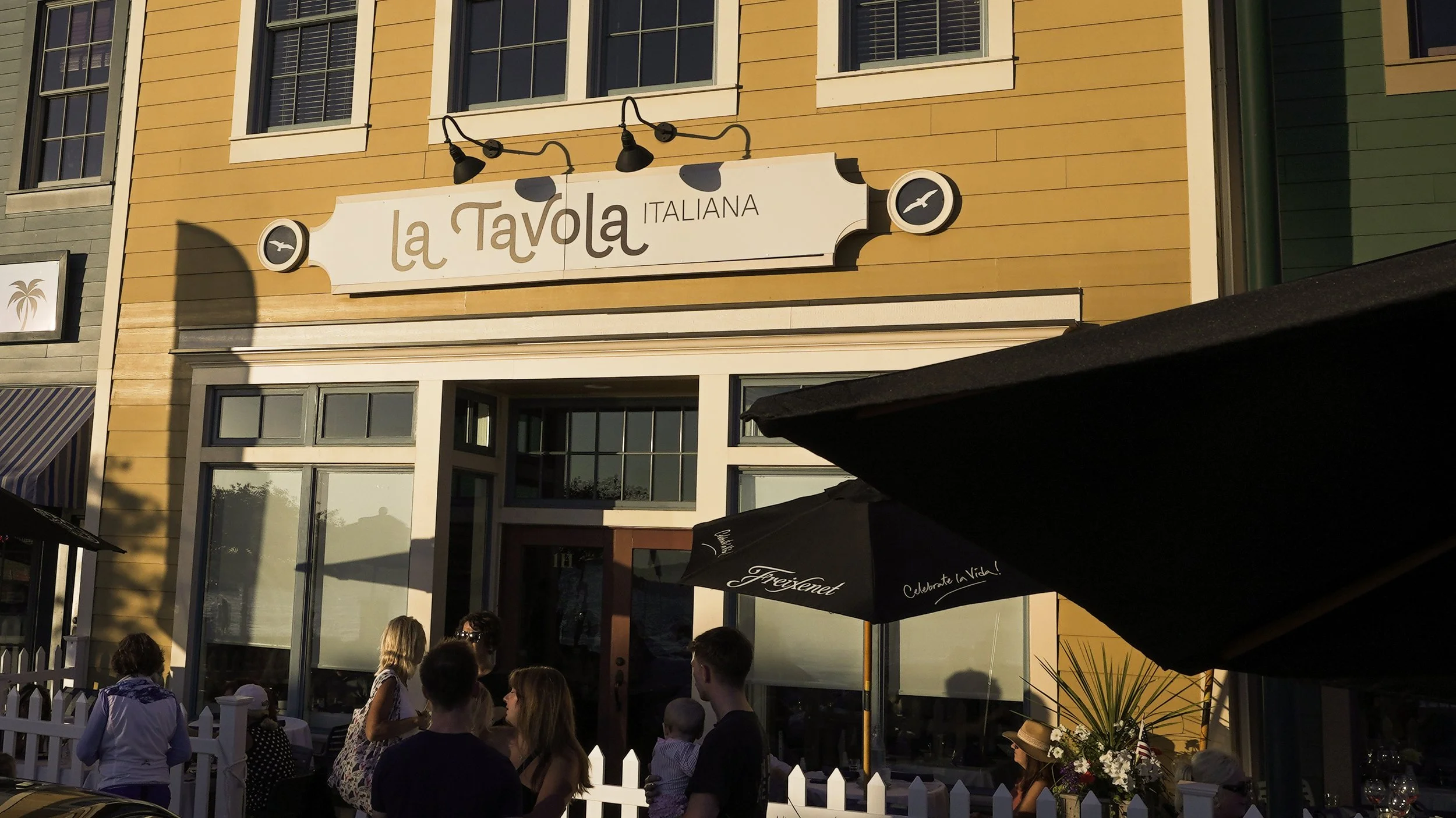 Exterior of a restaurant named La Tavola Italiana with people gathered outside, some standing and others sitting, during the day. The building has yellow siding with multiple windows, and part of a patio umbrella is visible in the foreground.