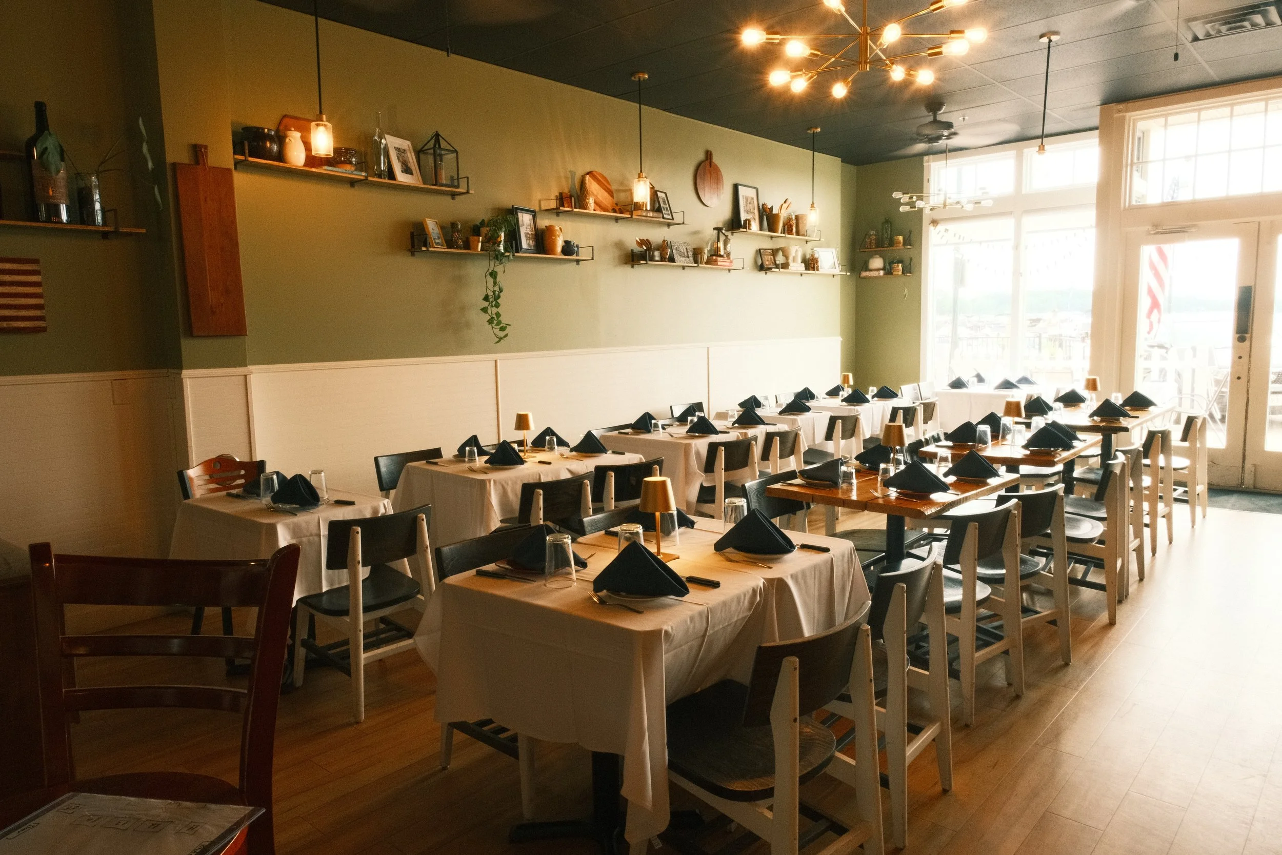Dining area in a restaurant with tables set with black napkins, glasses, and small lamps. The interior features a green wall with floating shelves holding decorative items, pictures, and plants. Large windows let in natural light.