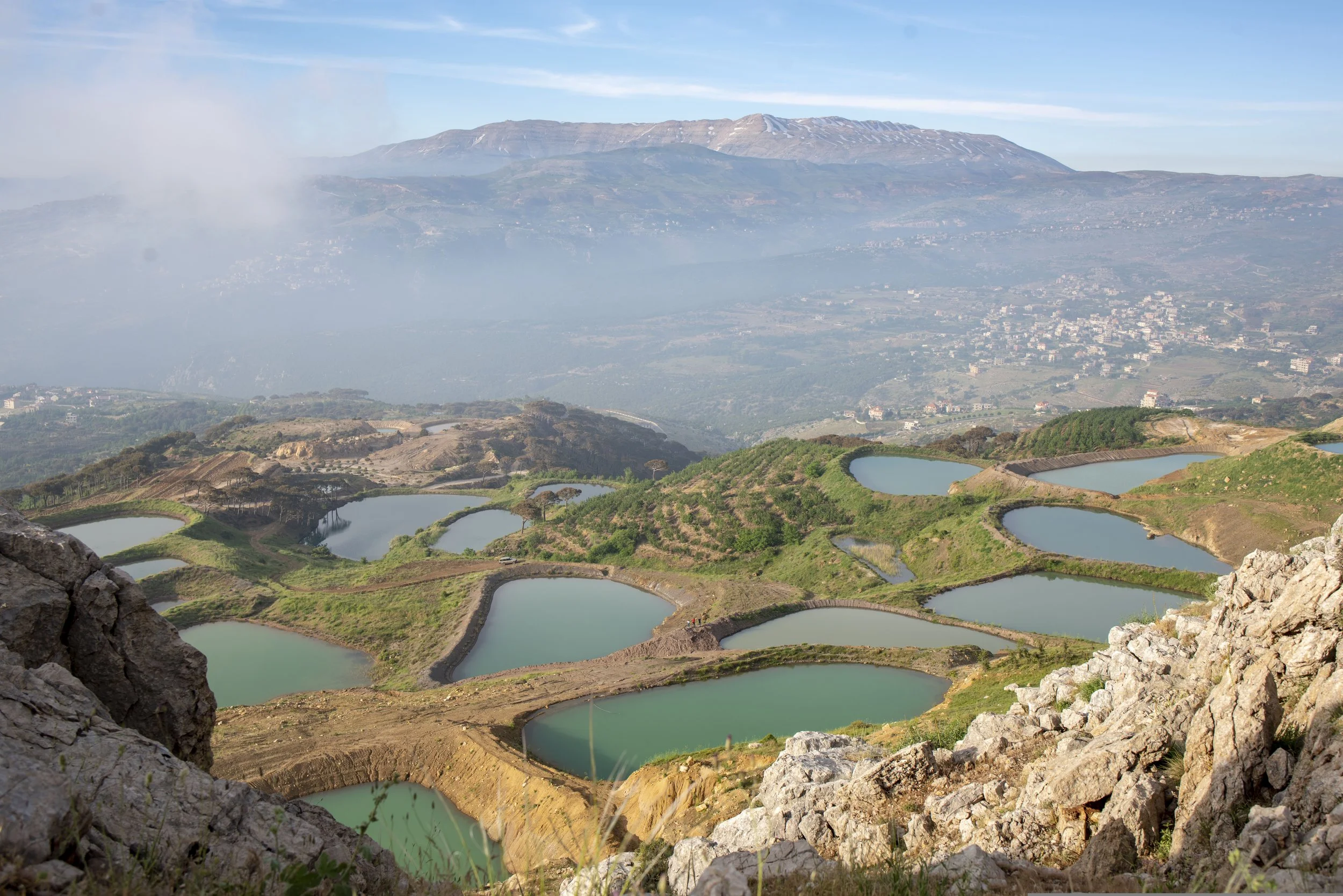 Mountain landscape with multiple small ponds in the foreground, scattered trees and shrubs, a city or town at the base of the mountain, and a large mountain range in the background under a clear blue sky.