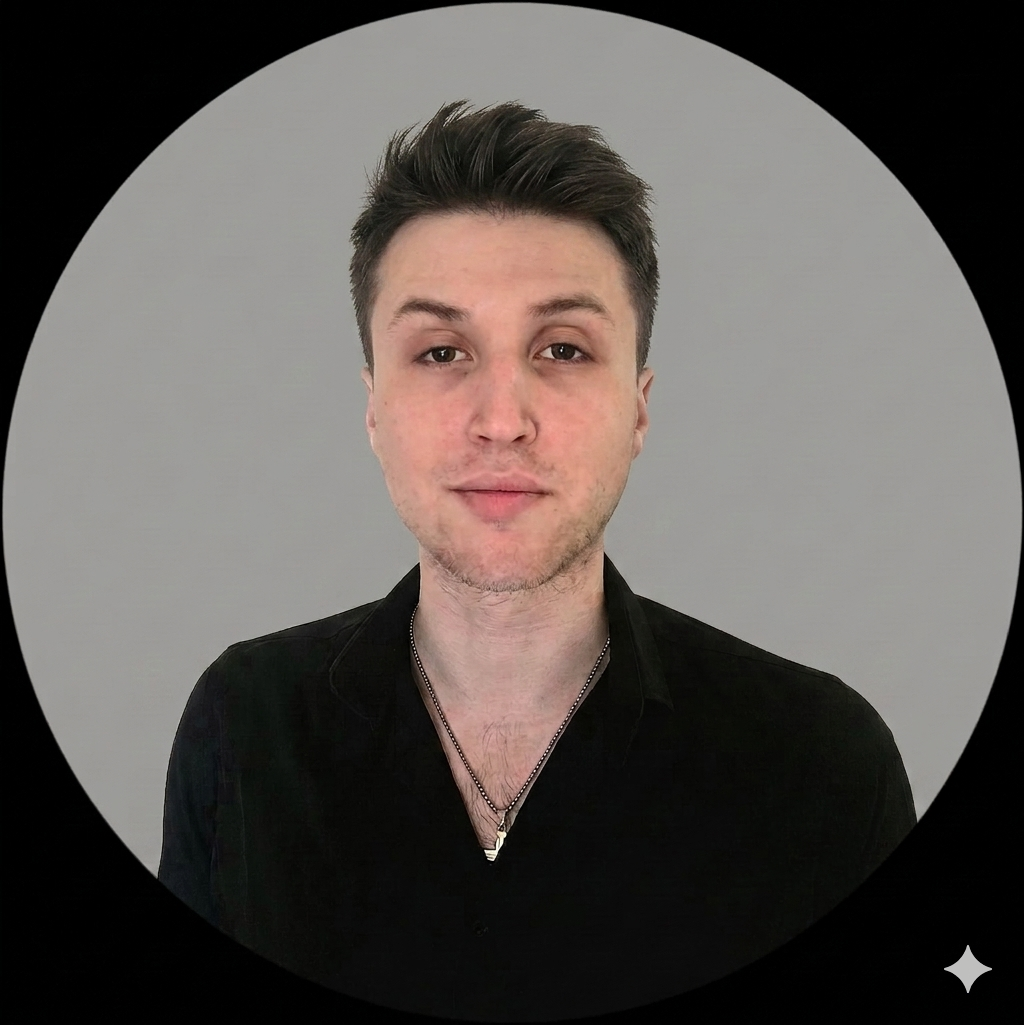 Portrait of a young man with short brown hair, fair skin, and wearing a black shirt with a silver chain necklace, standing against a light gray background.