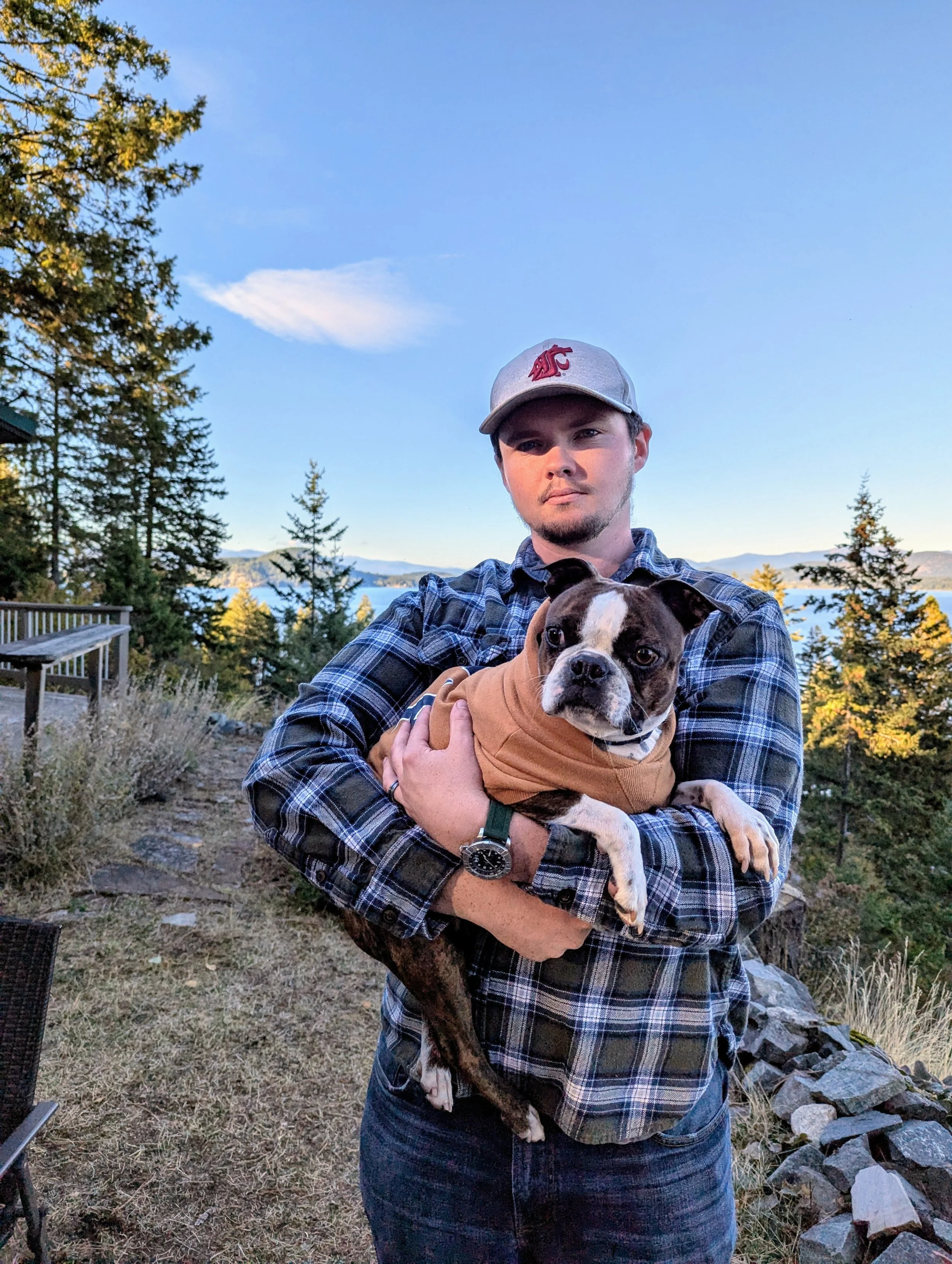 A young man wearing a grey baseball cap with a red logo, a blue plaid shirt, and a watch, holding a small dog with a brown jacket outdoors on a gravel path, with trees and mountains in the background during daytime.