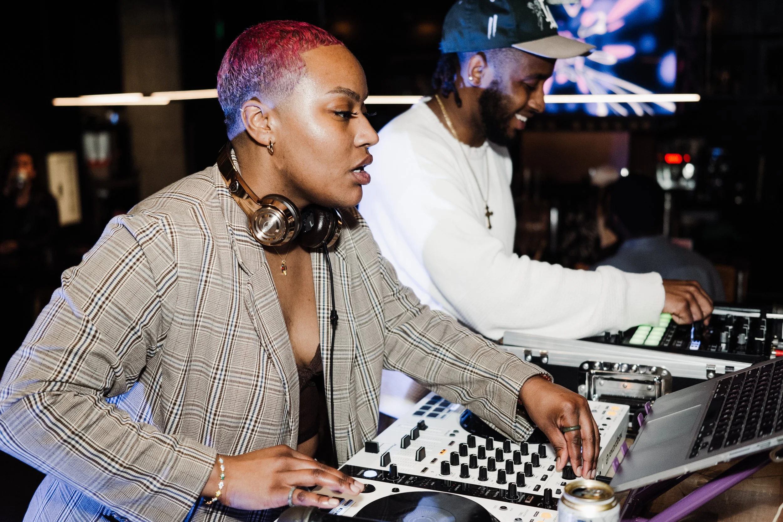 A woman with short pink hair and headphones around her neck is DJing at a nightclub, alongside a man in a baseball cap who is also DJing. They are focused on their equipment with colorful lighting in the background.