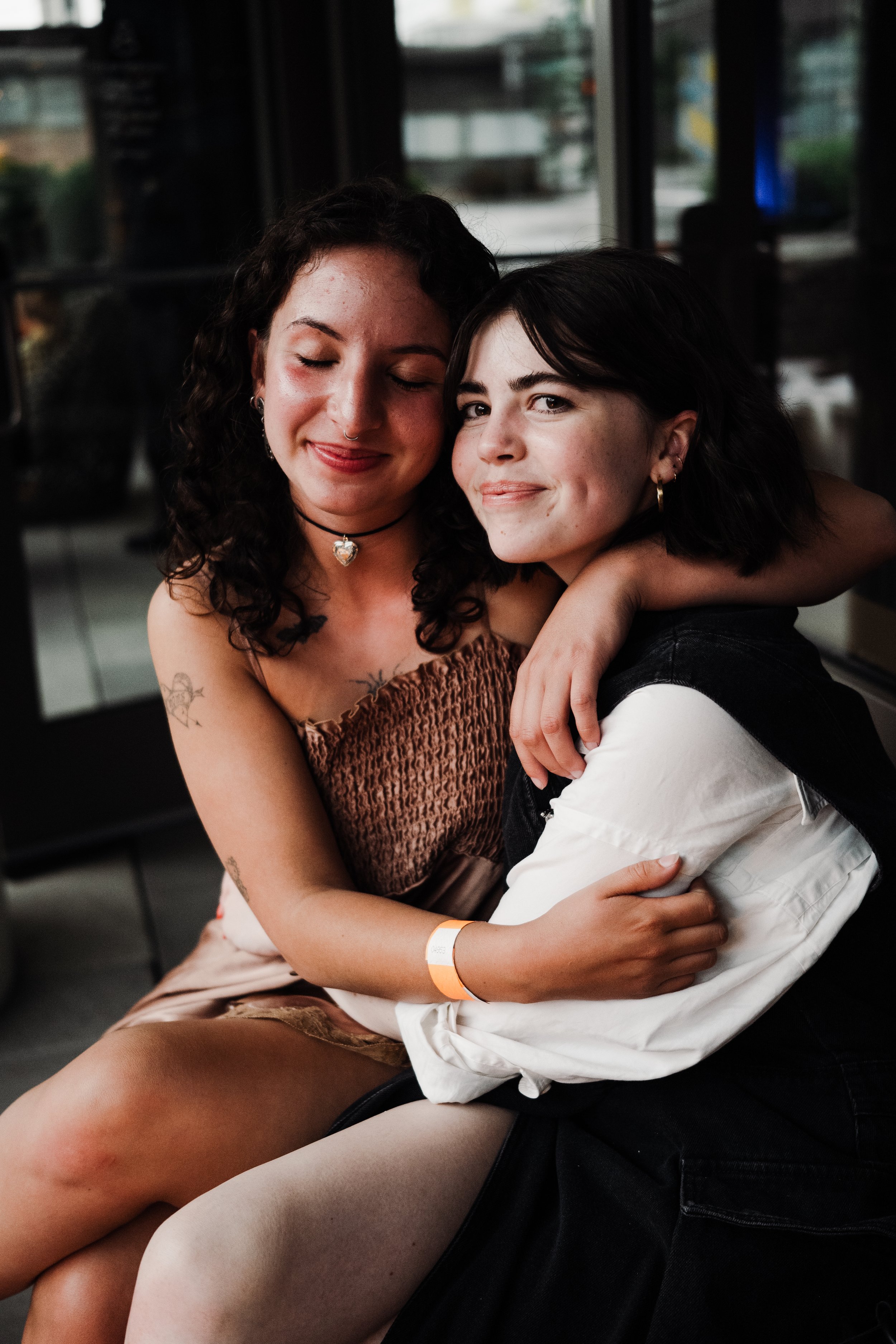 Two young women sitting close to each other, smiling and hugging, inside a dimly lit indoor space with windows in the background.