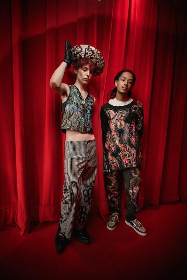 Two models dressed in colorful, patterned clothing stand against a red curtain backdrop. The model on the left wears a sleeveless top, striped pants, and a large hat, while the model on the right wears a long-sleeved, butterfly-printed shirt and matching pants.