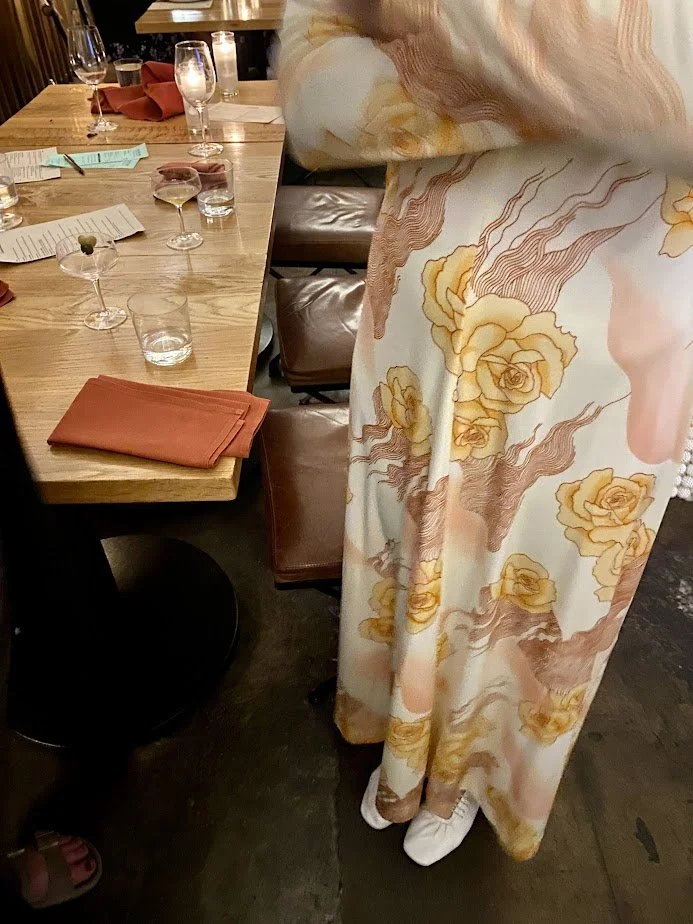 People dining at a restaurant table with glasses of wine and menus. A person is standing wearing a long, flowy dress with floral and wave patterns, paired with white shoes.