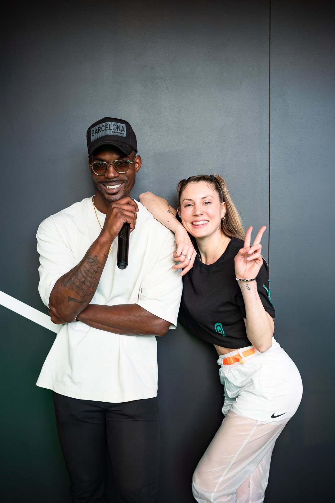 Two people smiling and posing for photo against a dark gray background. The man on the left is wearing glasses, a cap that says "Barcelona," and a white t-shirt, holding a microphone. The woman on the right is smiling, making a peace sign with her right hand, and has her left arm resting on the man's shoulder. She is wearing a black t-shirt and white pants.