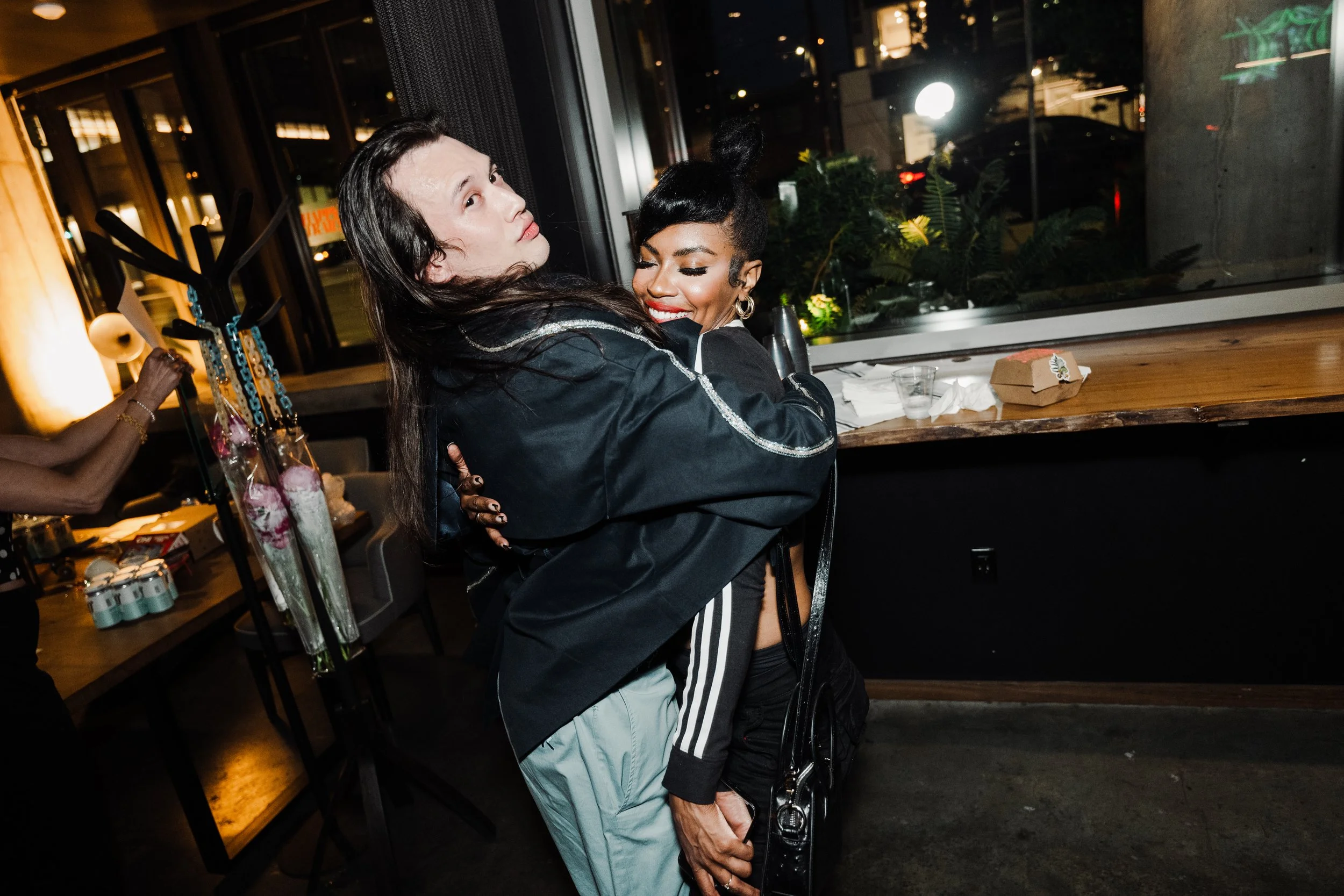Two  hugging each other in a dimly lit indoor space, possibly a bar or restaurant, with large windows showing a night scene outside. One woman has long dark hair, and the other has short black hair styled in a bun. There are drinks and snacks on the table nearby.