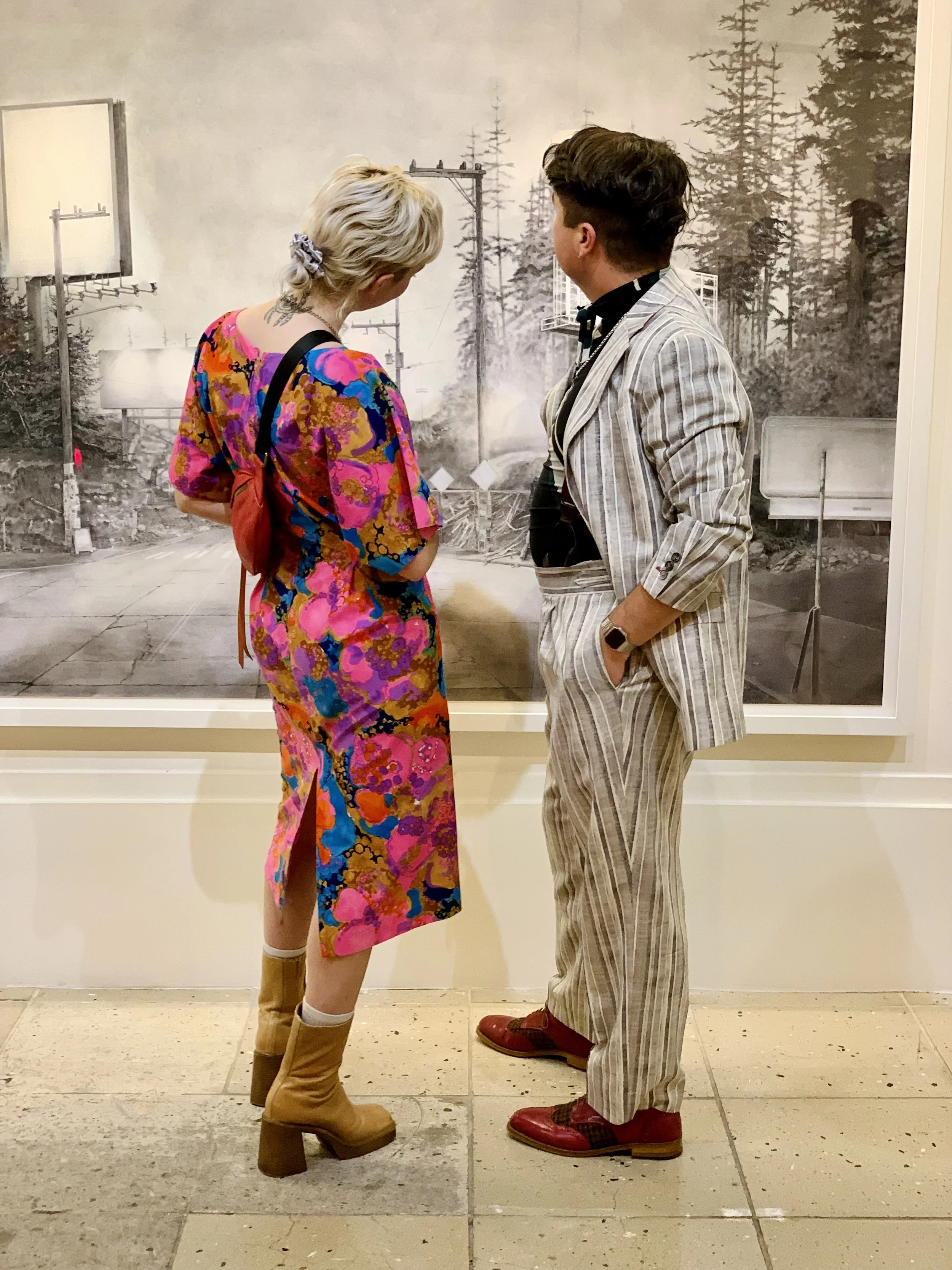 Two people are standing close and facing each other in an art gallery. The person on the left has short, blonde hair, a colorful floral dress, and tan boots. The person on the right has dark hair styled in a pompadour, a light striped suit with a black shirt, and red shoes. There is a large black and white forest landscape painting in the background.
