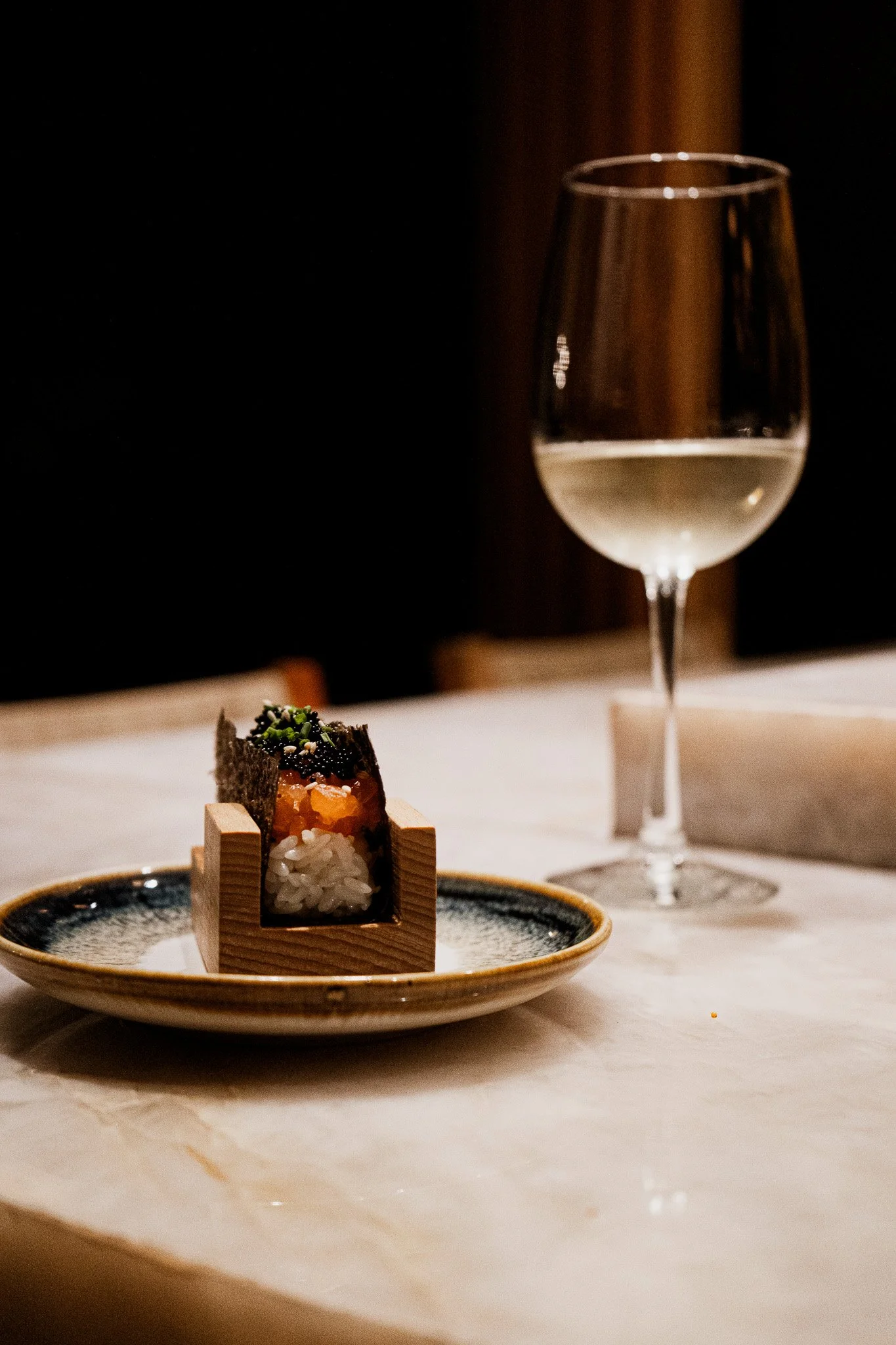 Vaso de vino blanco sobre mesa, con comida japonesa en plato, fondo oscuro.