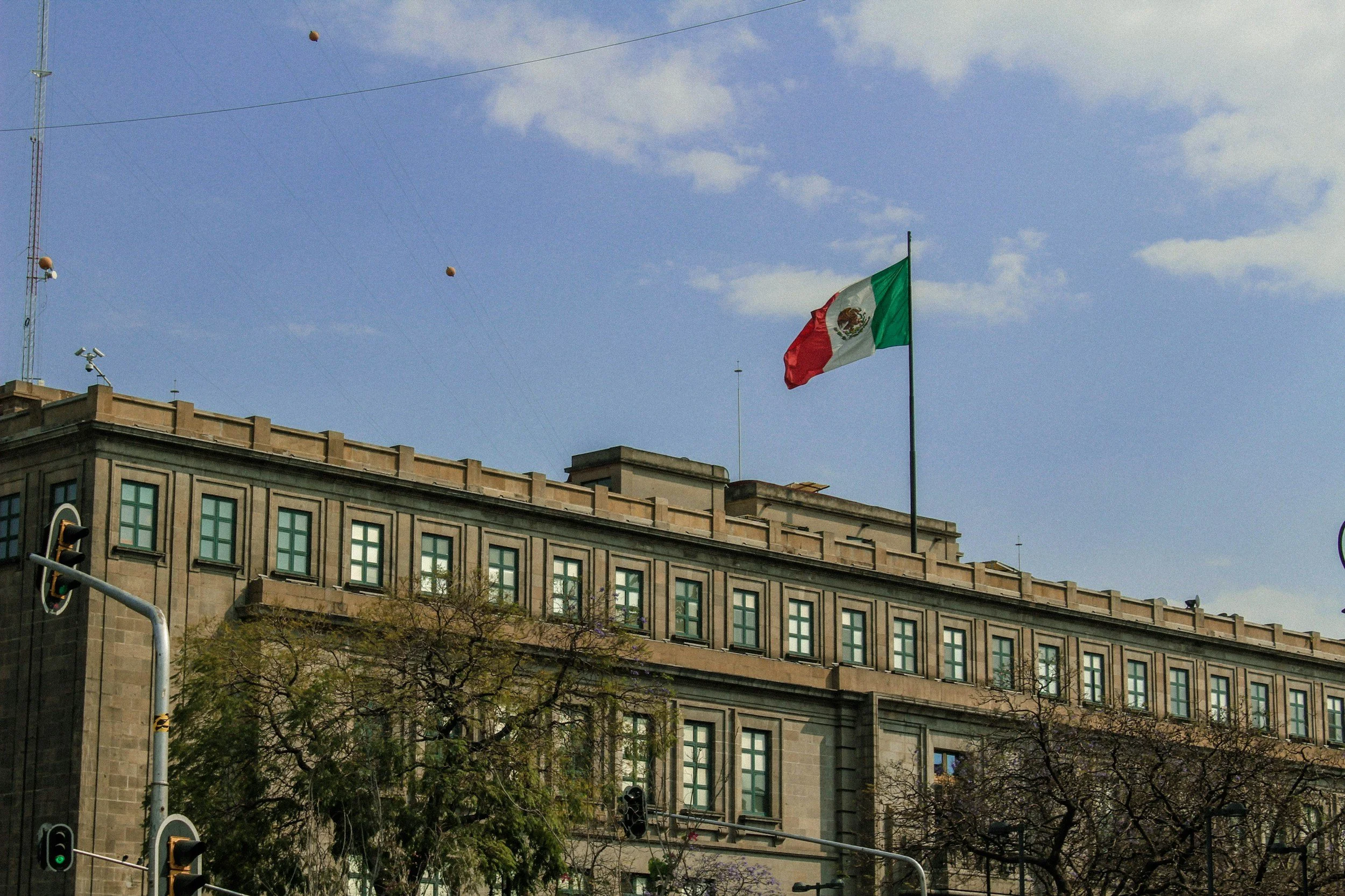 Edificio con bandera de México ondeando en la parte superior, cielo azul con algunas nubes, semáforo en primer plano y árboles en el lugar.