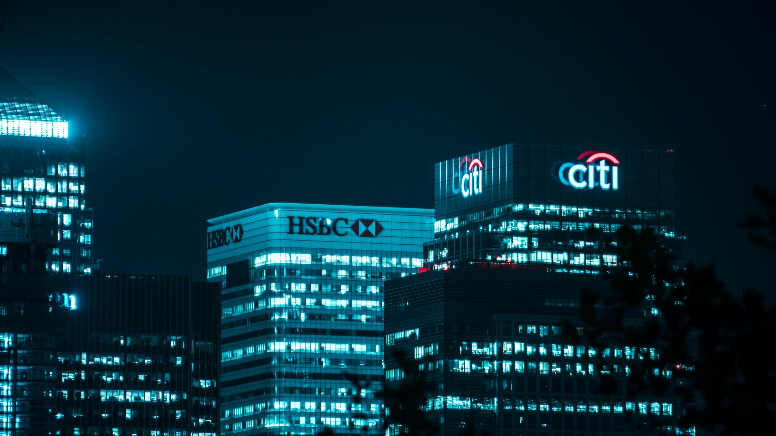 Edificios de oficinas en una ciudad de noche con luces neón, mostrando los logotipos del banco Citi, HSBC y otros bancos en los rascacielos.