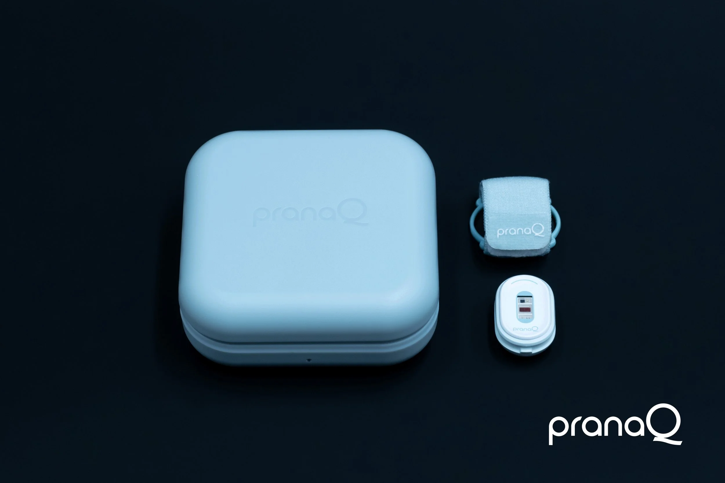A set of blue and white PranaQ sleep products on a dark surface, including a rectangular device, a small strap, a clip with a display, and a small round object.