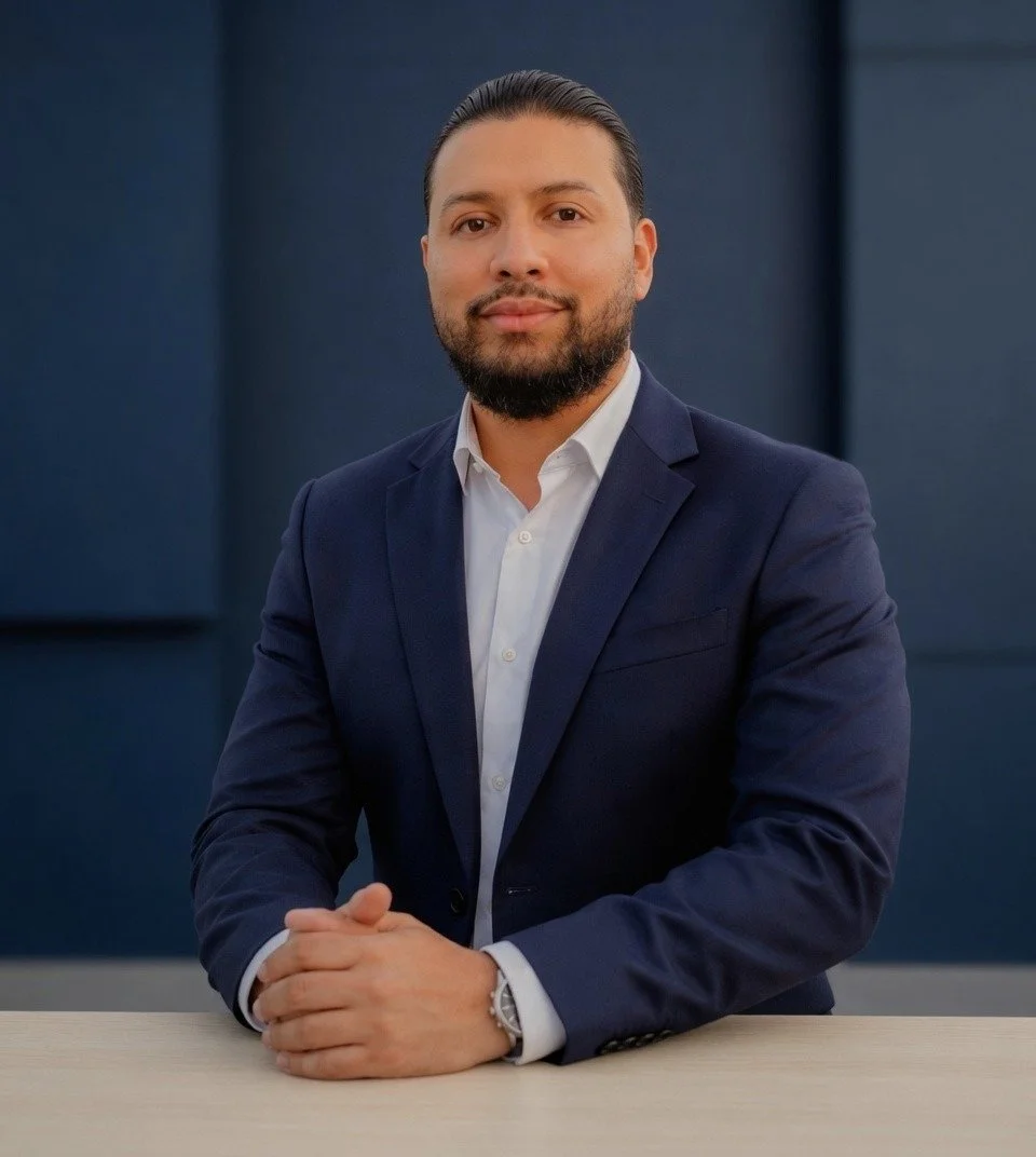 A man with dark hair, beard, and light skin wearing a dark blue suit and white dress shirt sitting at a light-colored table with hands clasped, against a dark blue background.