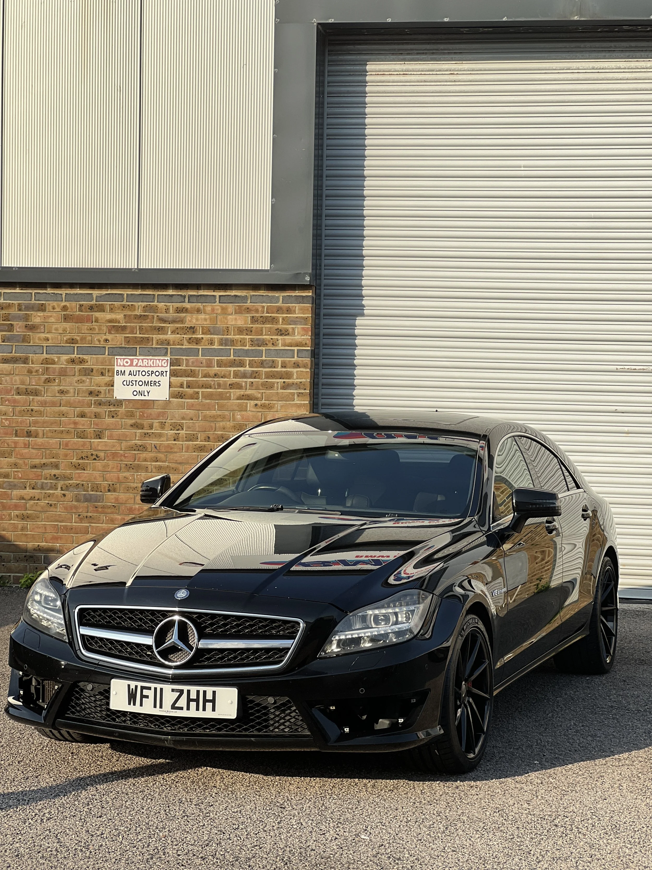 Black Mercedes-Benz car parked in front of a brick wall and metal building with a roller shutter door, with a sign that reads 'No Parking, BM Autosport, Customers Only'.