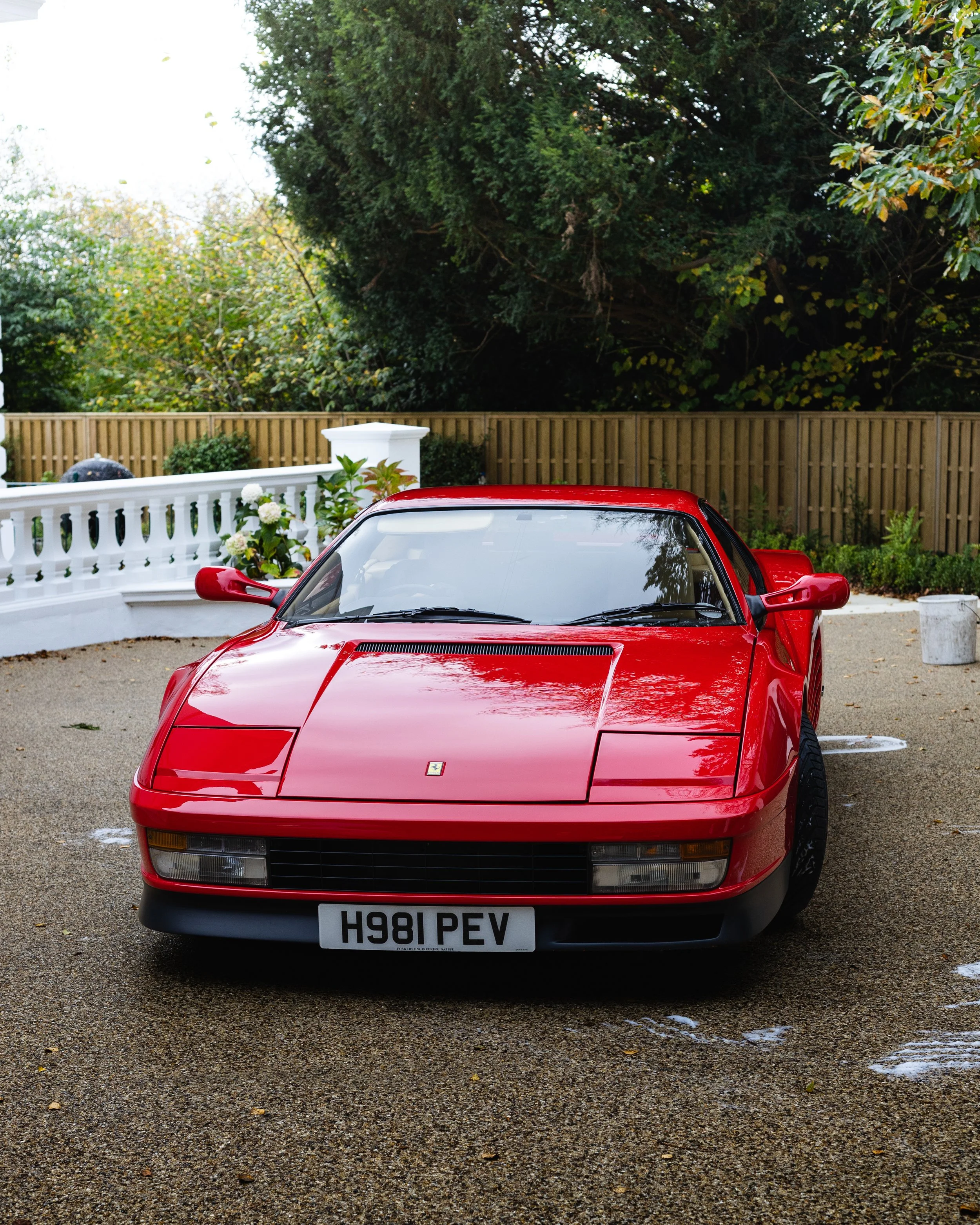 Red vintage Ferrari sports car parked on a gravel driveway with a white fence and greenery in the background.