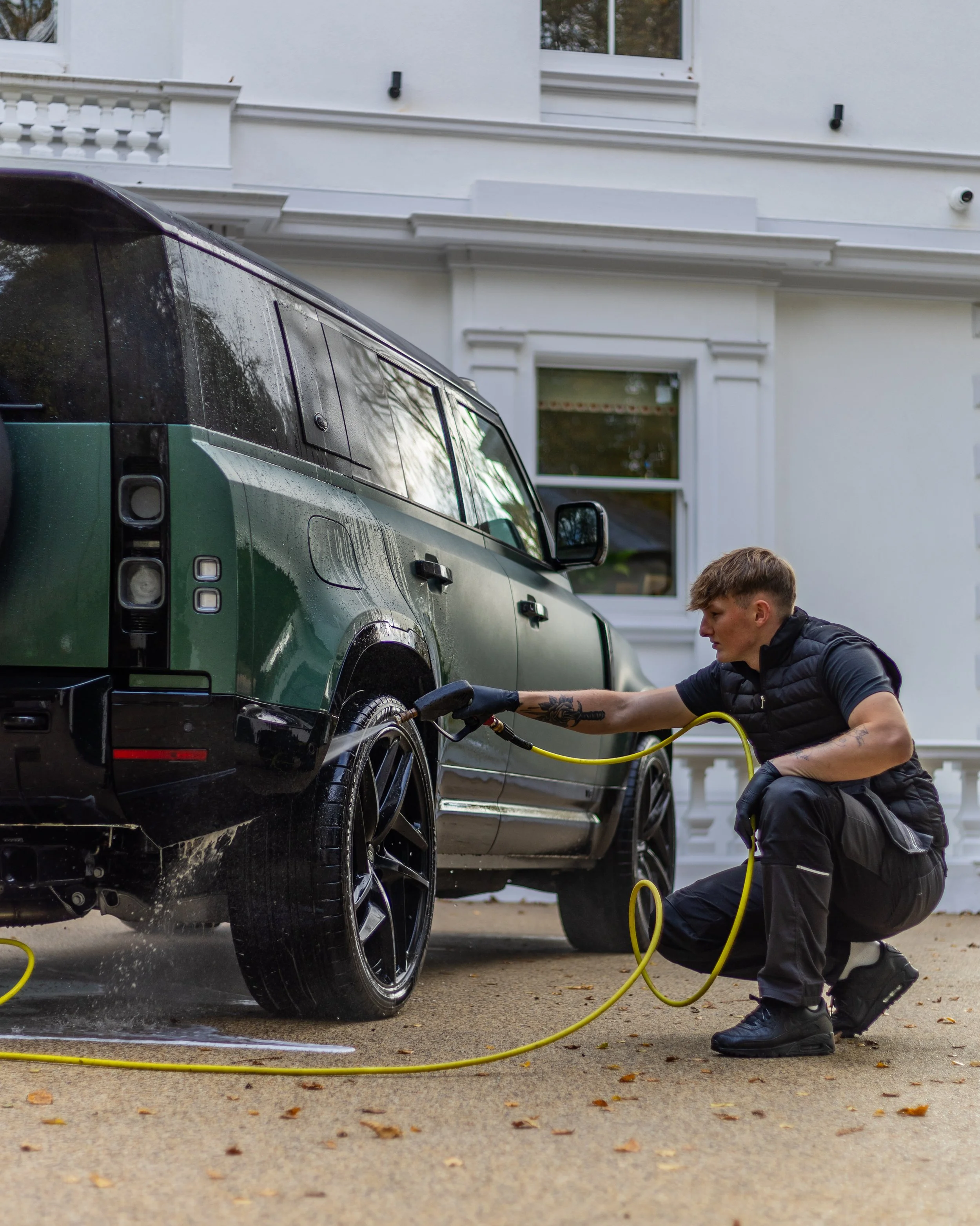 Person washing a black and green SUV with a pressure washer in front of a white house.