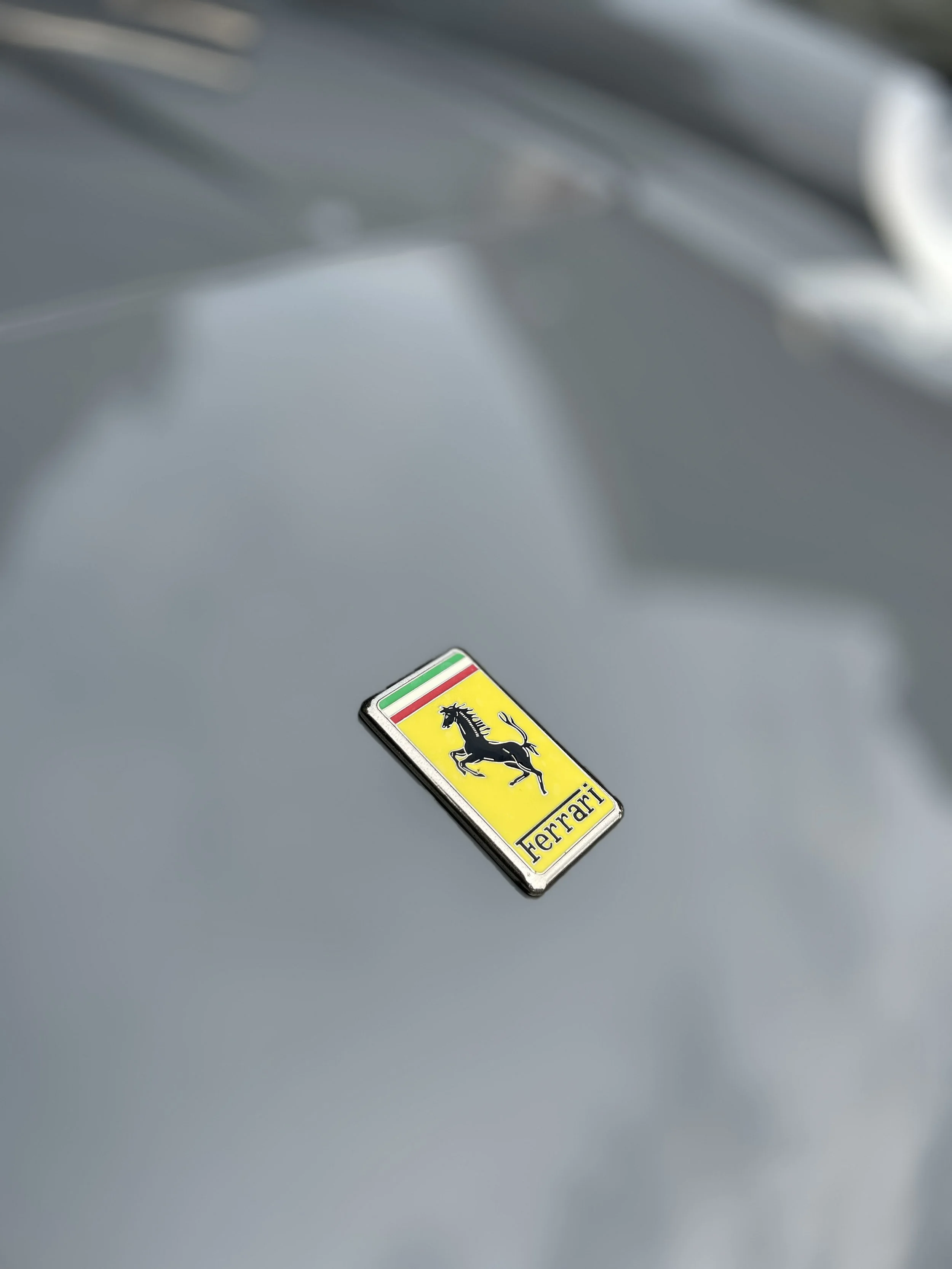 Close-up of a Ferrari emblem with a black prancing horse on a yellow background and Italian flag stripes above, mounted on a silver surface.