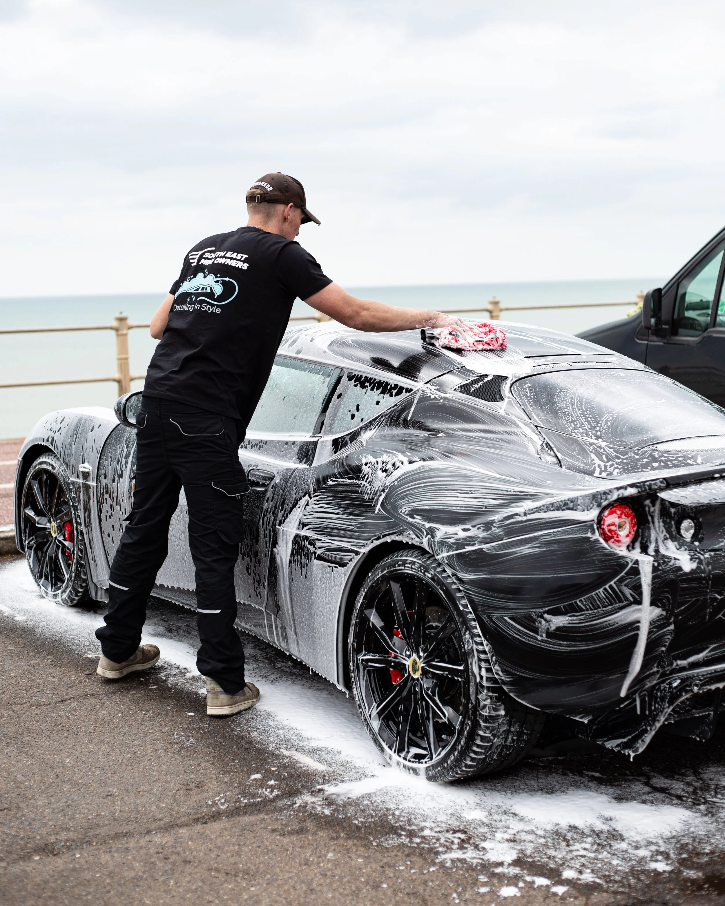Person washing a black sports car with soap and foam, near a beach.