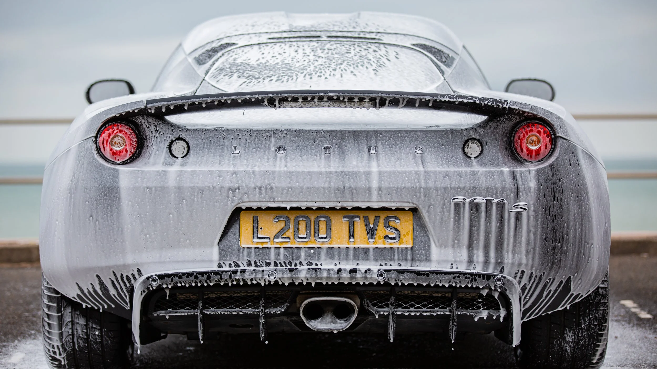 The rear view of a silver Lotus sports car covered in soap and water, with a license plate reading 'L200 TVS' and a cloudy sky in the background.
