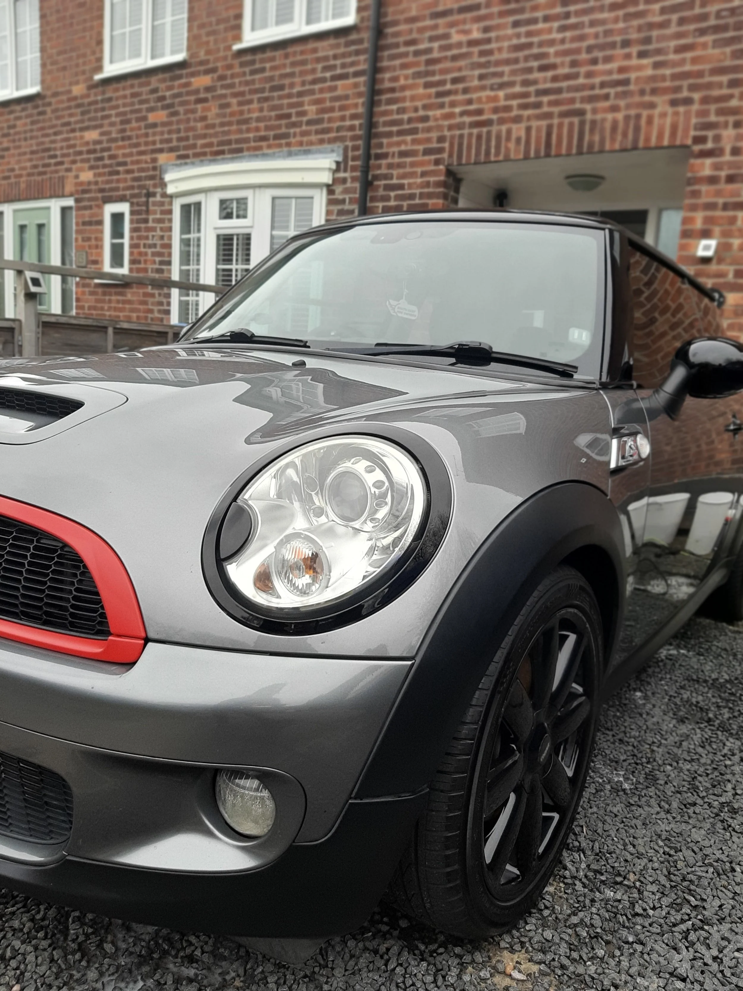 A gray Mini Cooper with black accents parked on gravel in front of a brick building.