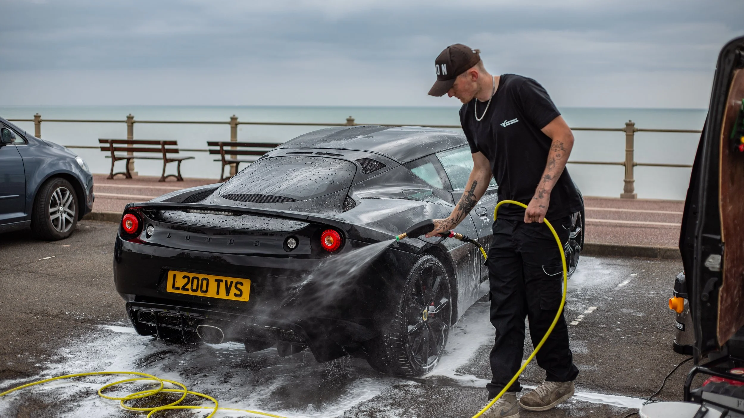 Man washing a black Lotus sports car with a hose at a seaside parking lot, with another car parked nearby and benches in the background.