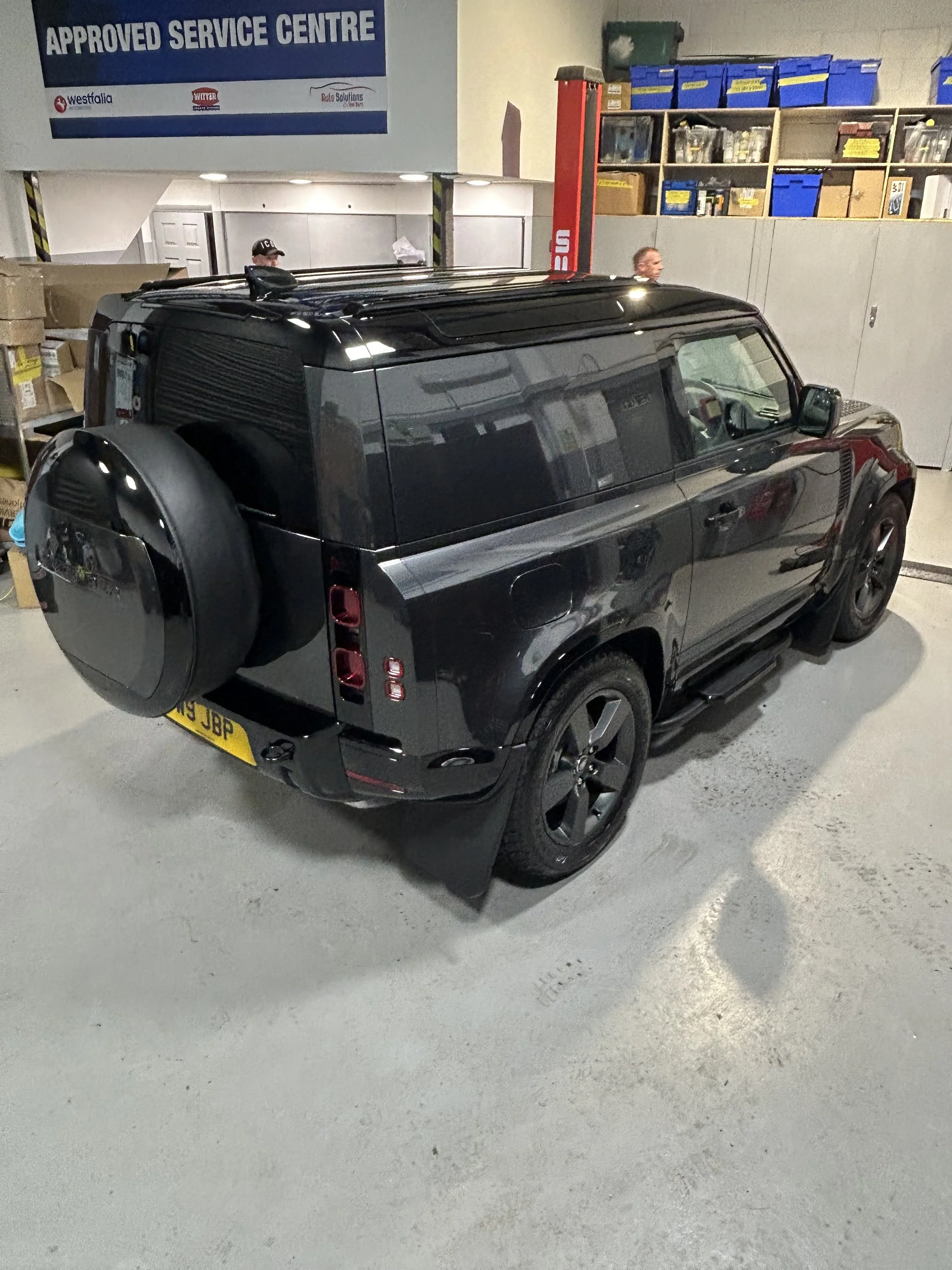 A black Land Rover Defender parked in an indoor garage near an approved service center sign, with a spare tire mounted on the rear.