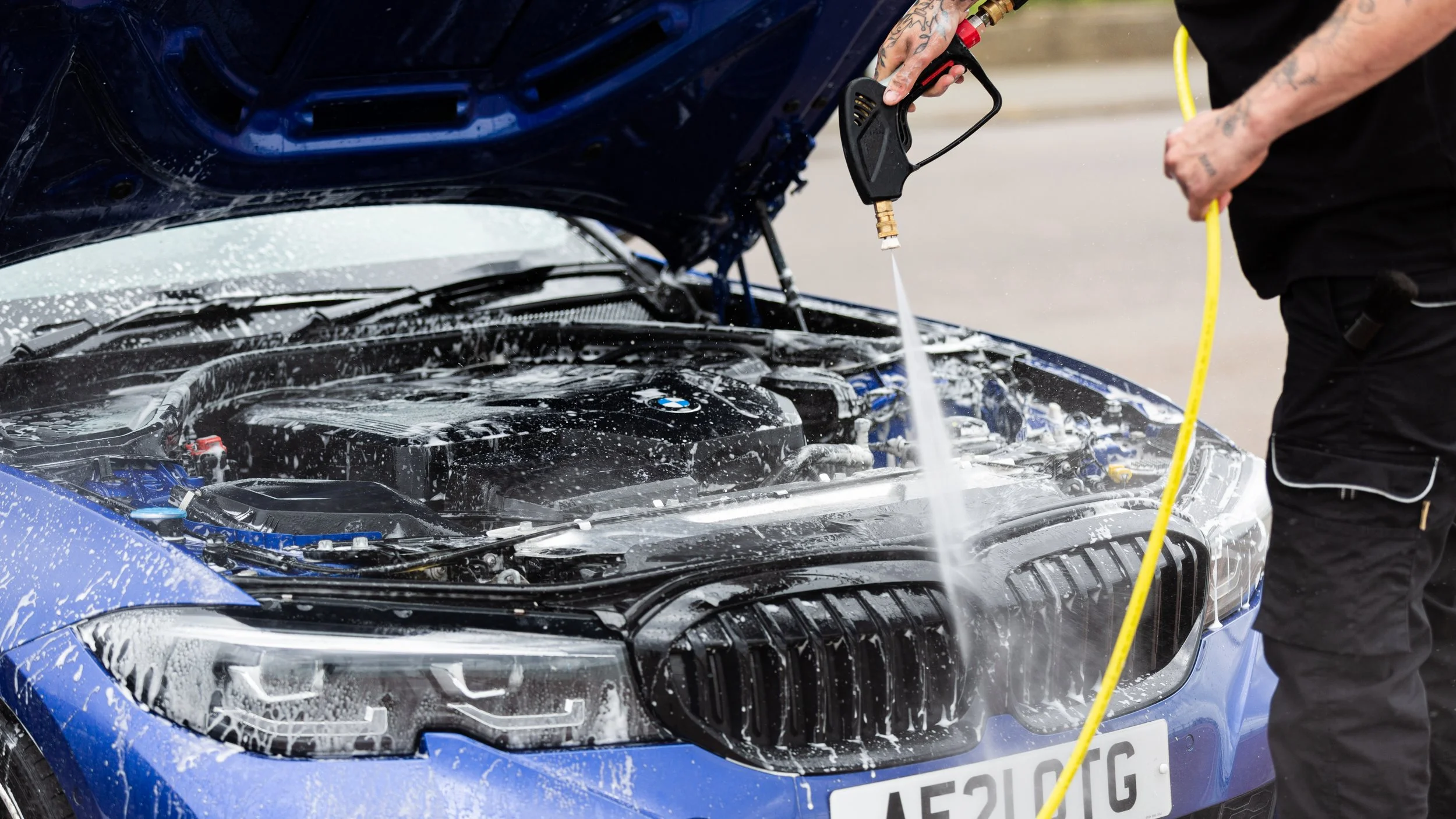 Person washing a blue car with a hose and soap under the open hood.