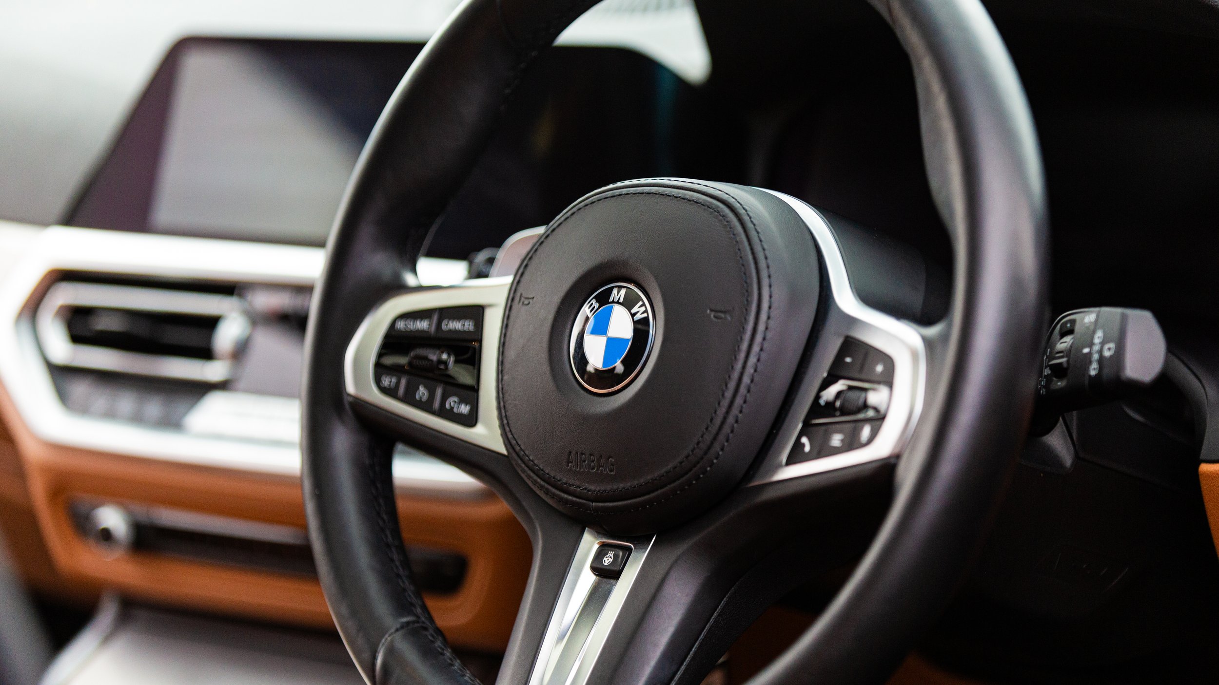 Close-up of a BMW steering wheel with controls, interior dashboard with air vents and display in the background.