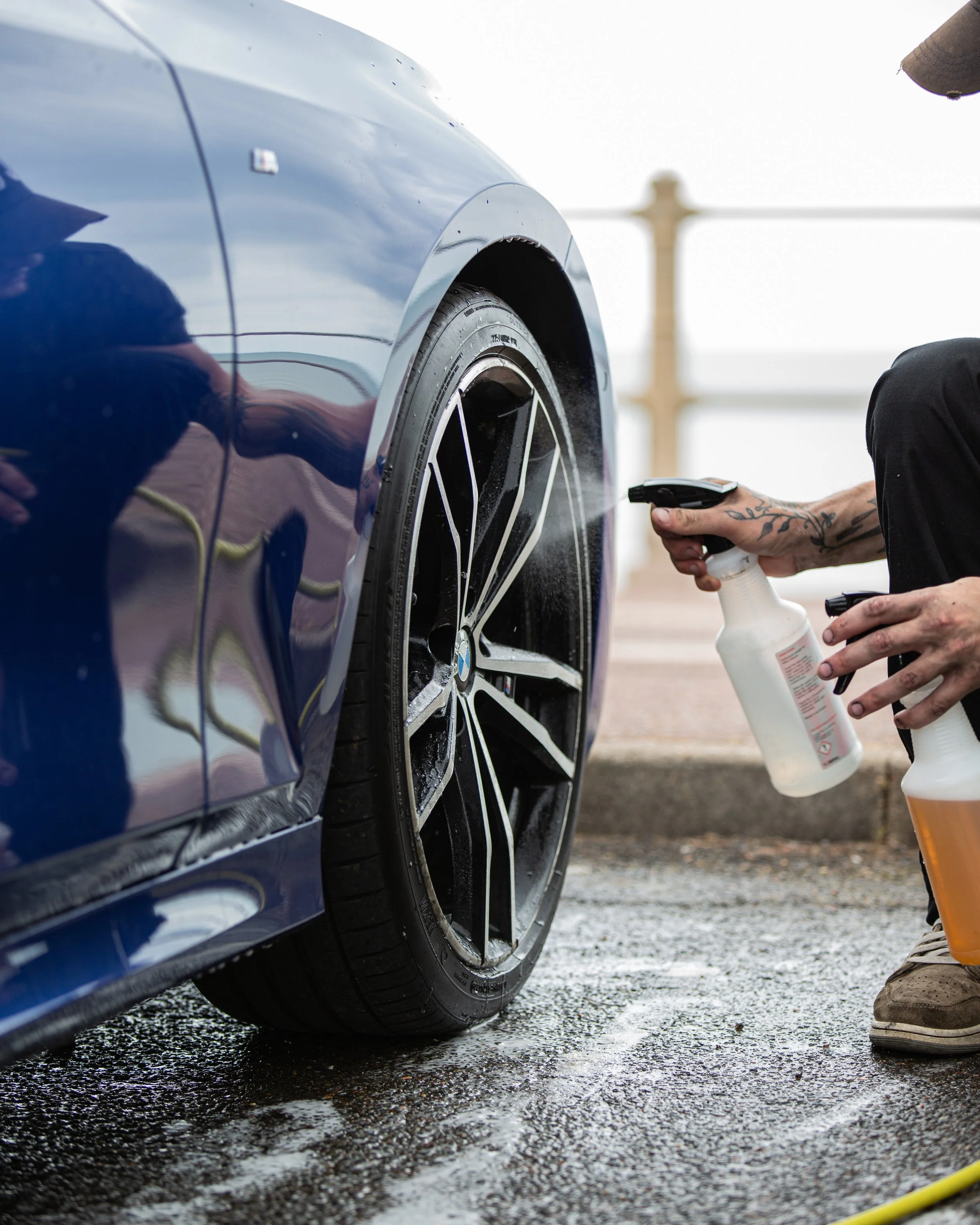 Person cleaning a BMW car's wheel with spray bottles.