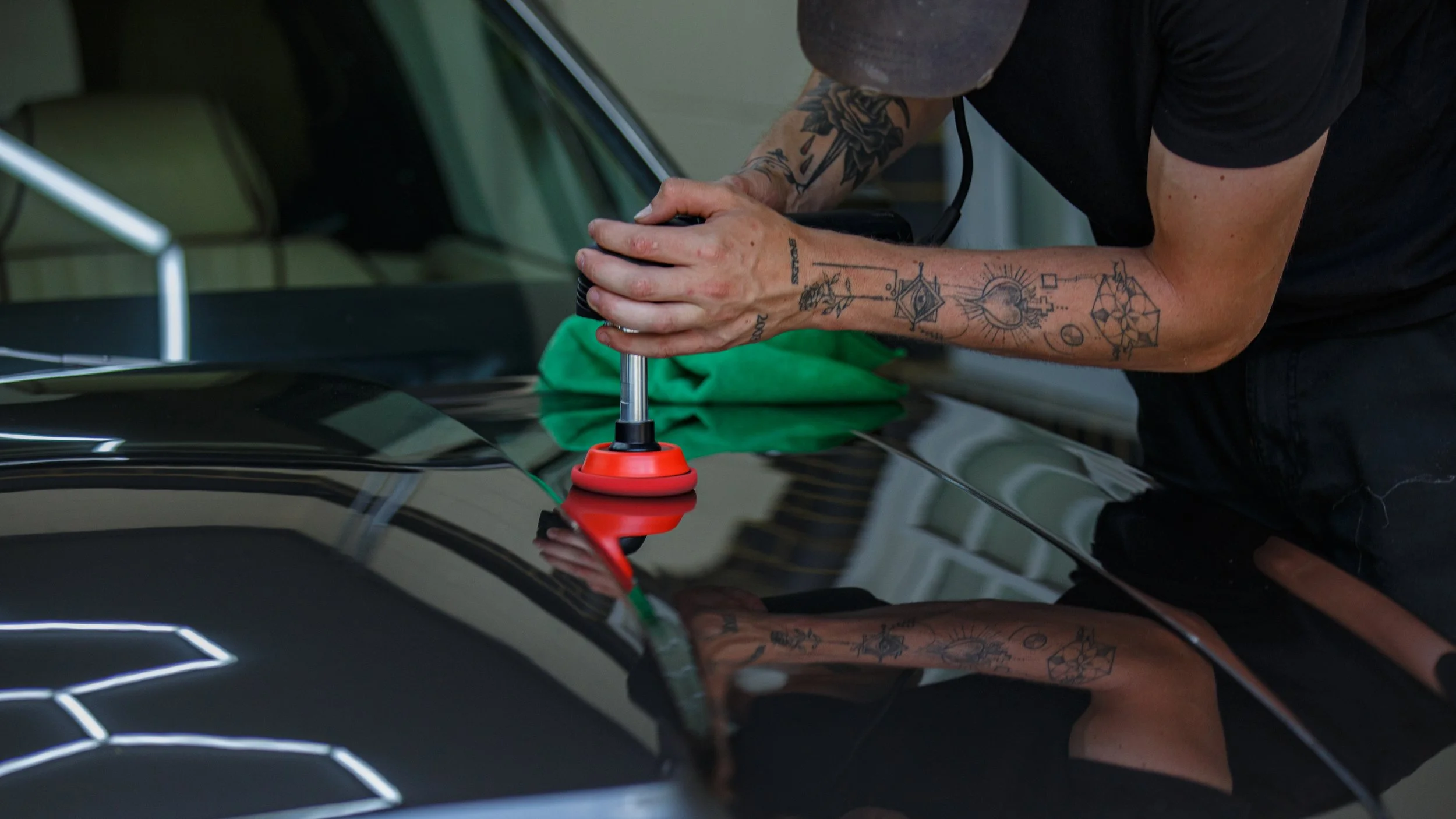 A person with tattoos on their arms uses a machine to polish or repair the surface of a black car.