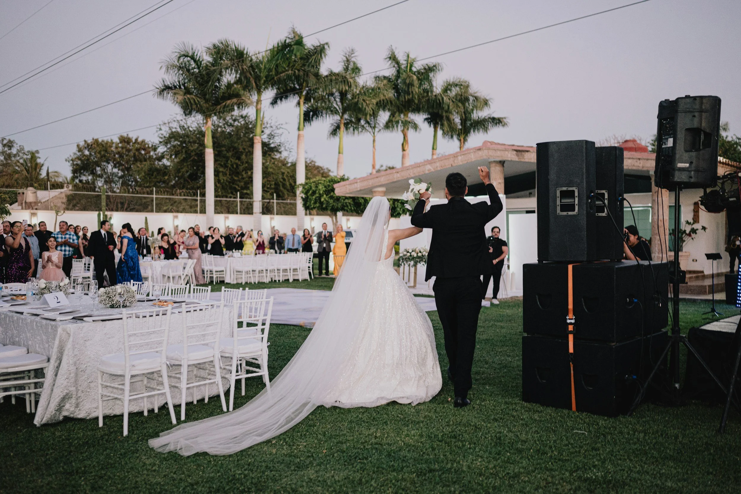 A bride and groom dancing at an outdoor wedding reception with guests watching. The bride wears a white wedding dress with a long train, and the groom wears a black suit. There are white chairs, tables, and floral centerpieces, and a backdrop of palm trees and a building, with speakers and musical equipment.