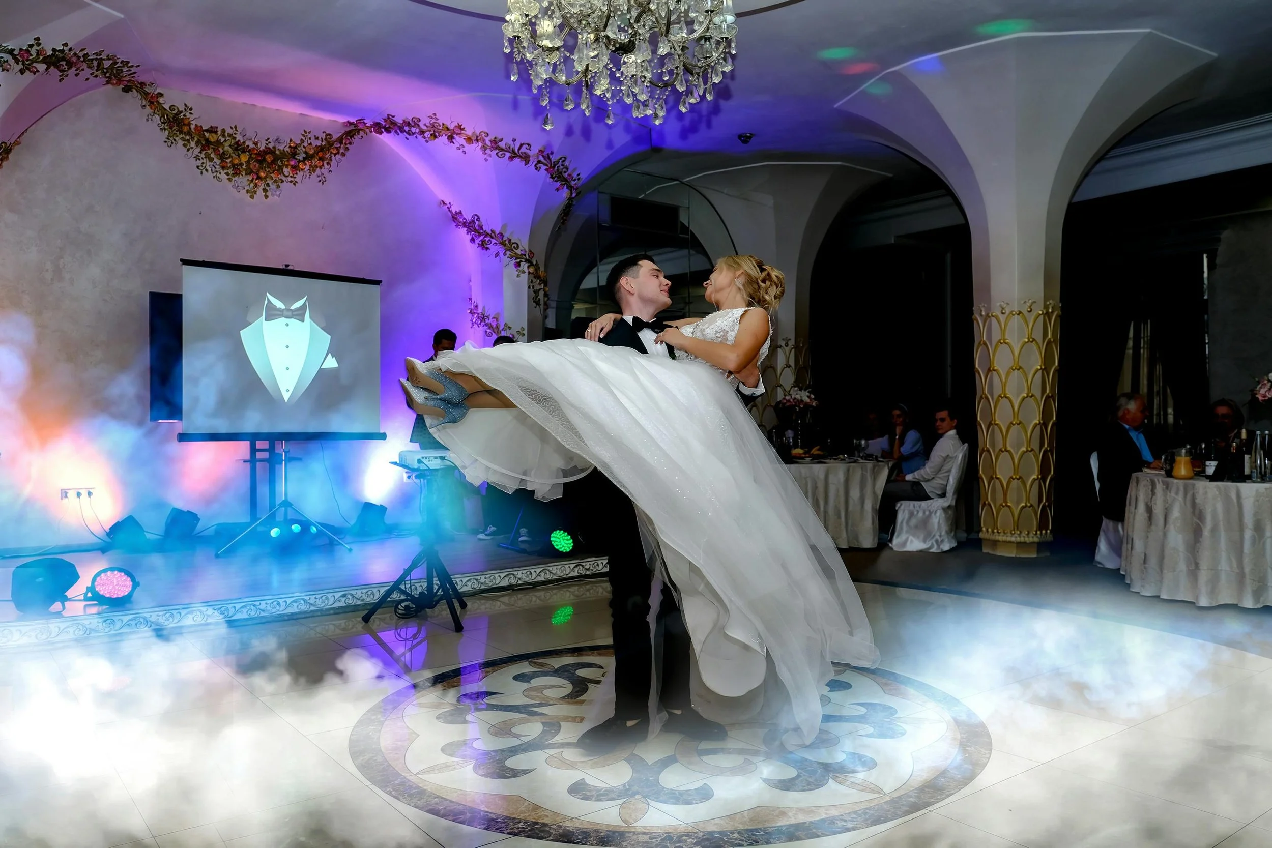 A bride and groom dancing at their wedding reception, with the groom lifting the bride in his arms. The venue has elegant arched ceilings, floral decorations, and a chandelier. There is a projection screen displaying a tuxedo graphic.