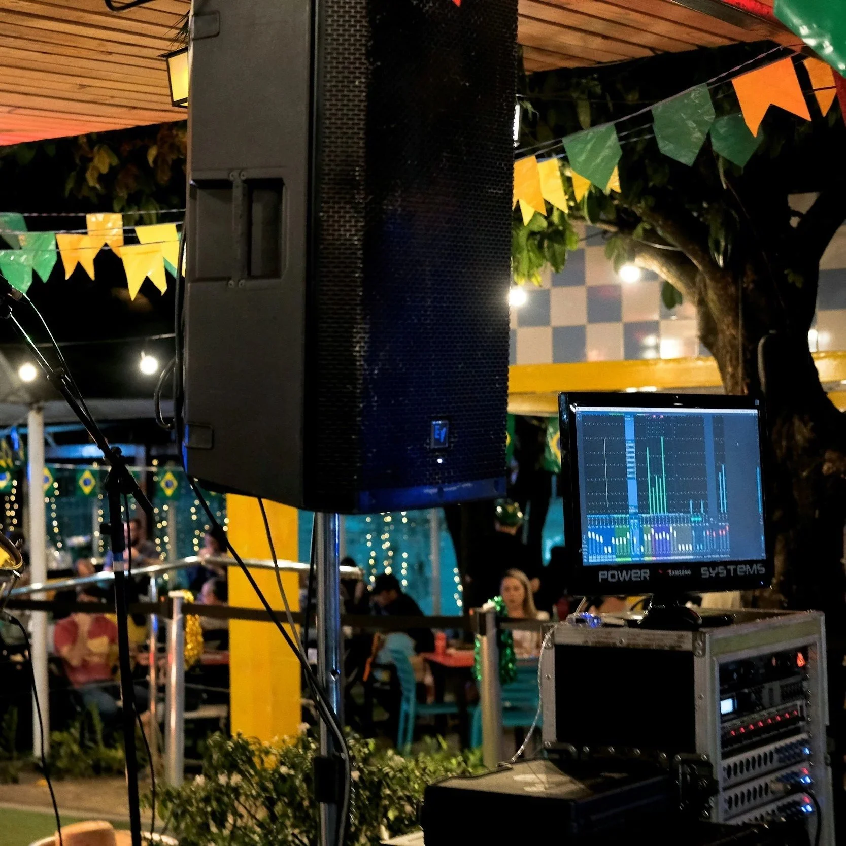 A DJ setup with a large black speaker, sound mixer, and a laptop displaying audio software at an outdoor event night. Decorations include green, yellow, and orange pennant banners, and string lights can be seen in the background.