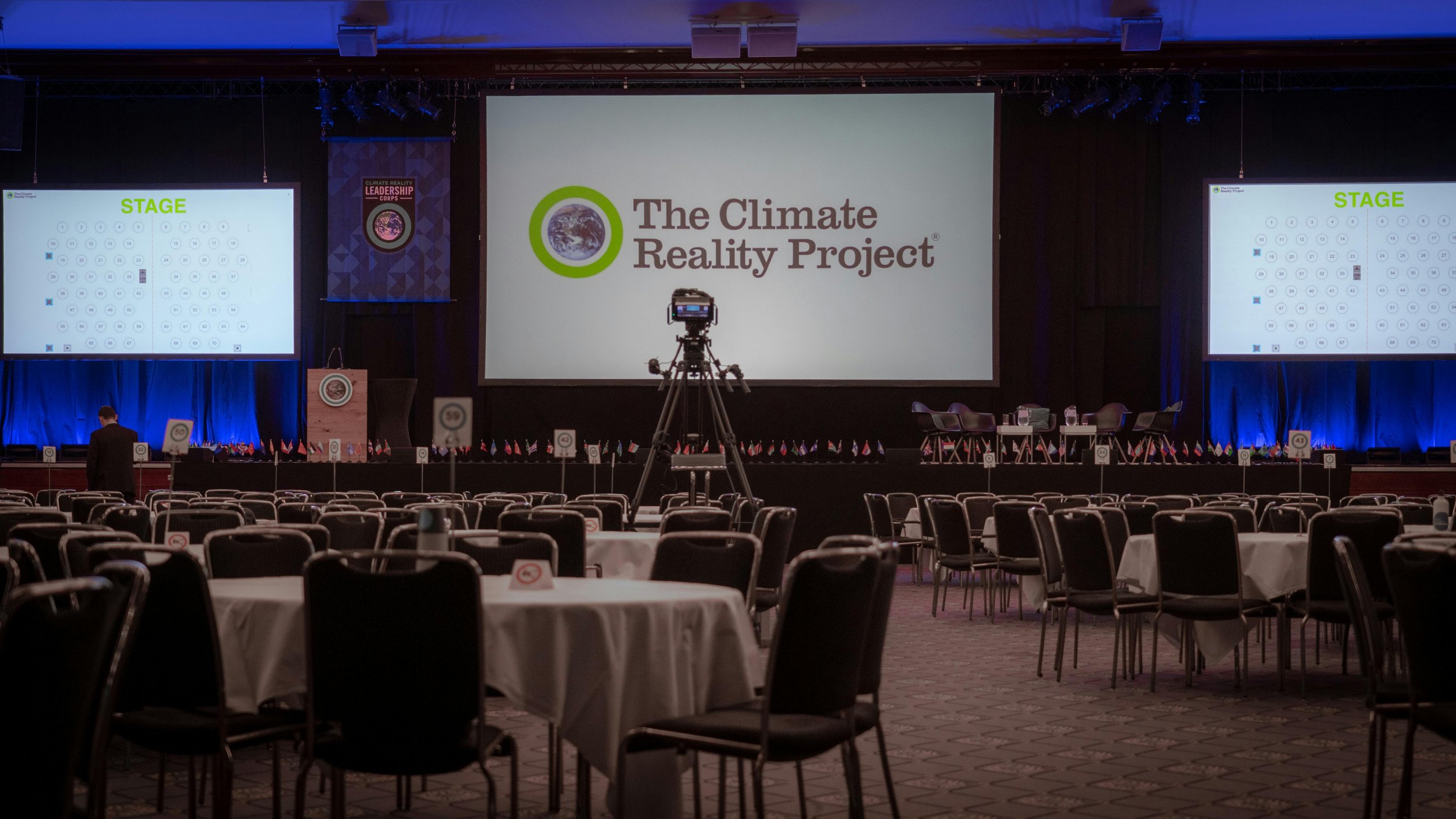 An empty conference hall with rows of tables and chairs, a large screen displaying 'The Climate Reality Project' logo, and two smaller side screens showing seating arrangements.