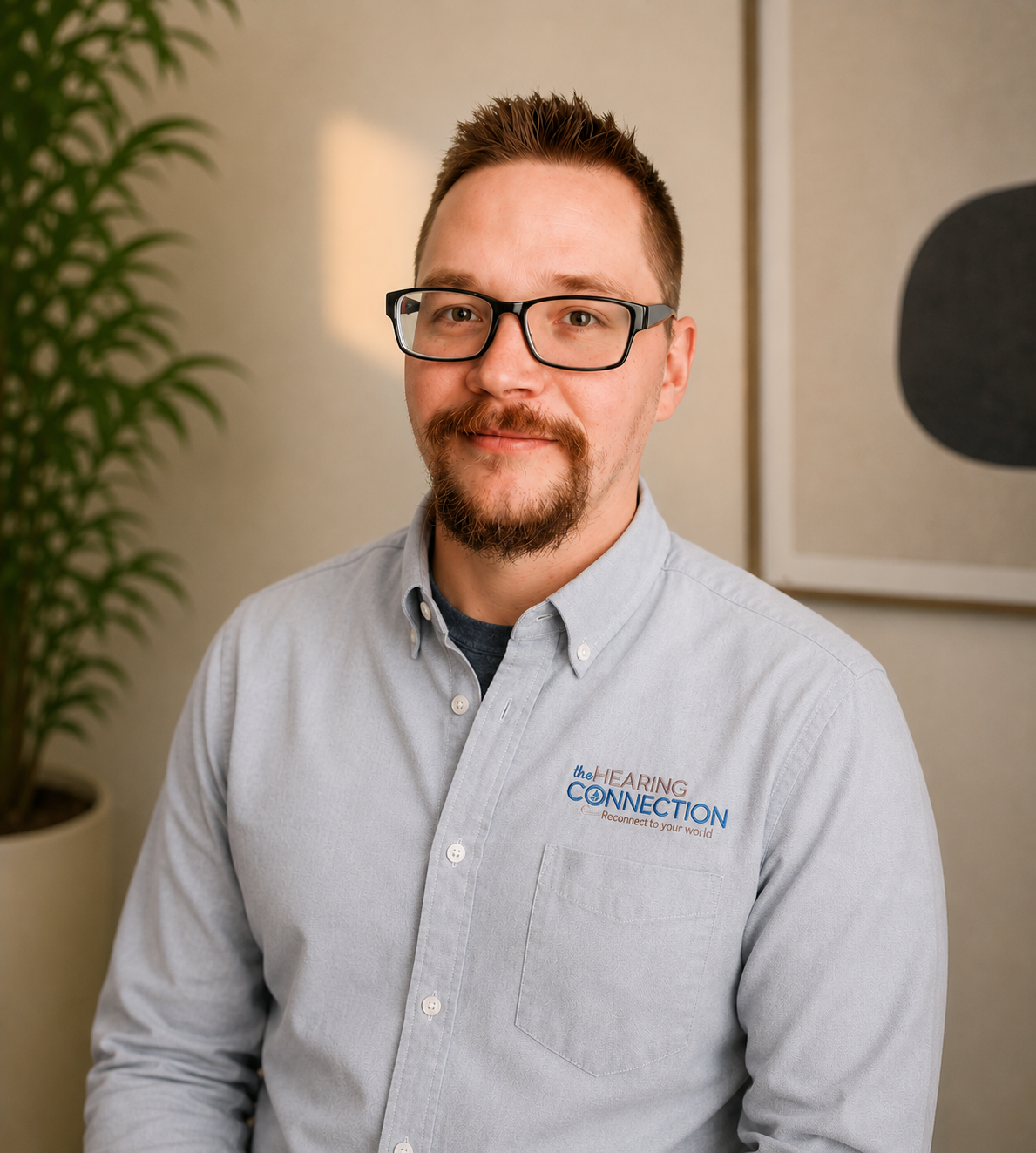 A man with glasses and a goatee, wearing a light gray button-up shirt with a logo that says "Hearing Connection," sitting indoors with a potted plant and abstract wall art in the background.