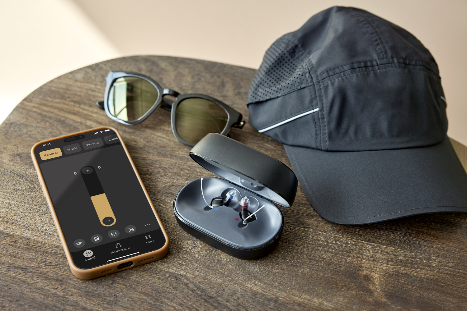 A wooden table with a pair of sunglasses, a black cap, a hearing aid case with hearing aids, and a smartphone displaying hearing aid controls.
