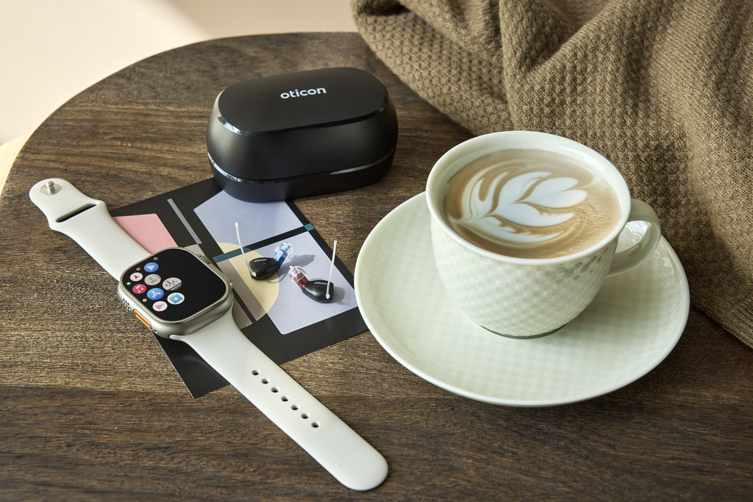 A wooden table holding a cup of coffee with latte art, a smartwatch, a black case with Oticon hearing aids, and a black charging case for hearing aids, with a part of a person's arm in a beige sweater in the background.