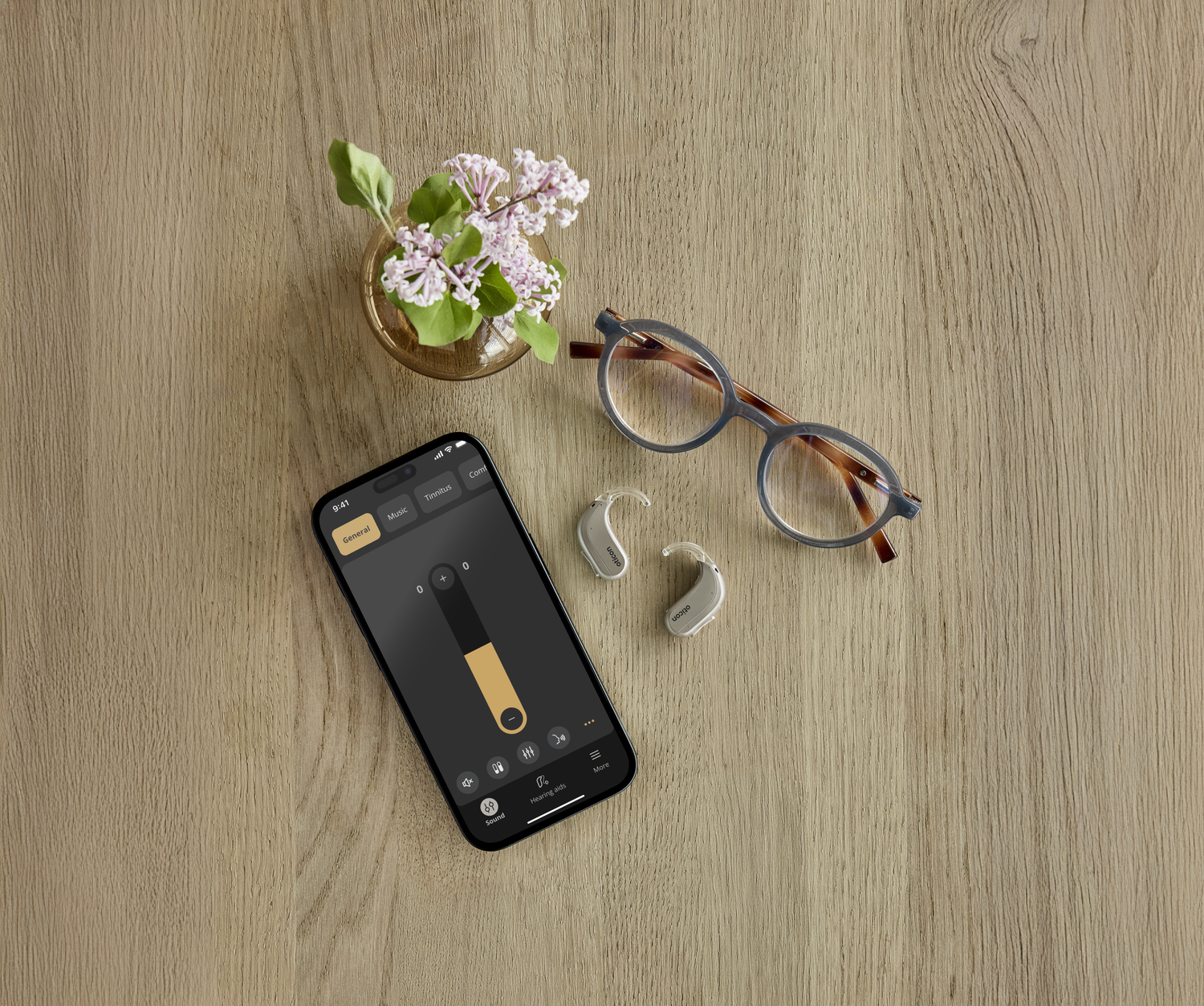 Wooden table with a smartphone displaying a sound control app, a pair of glasses, a hearing aid, a vase with pink flowers and green leaves.